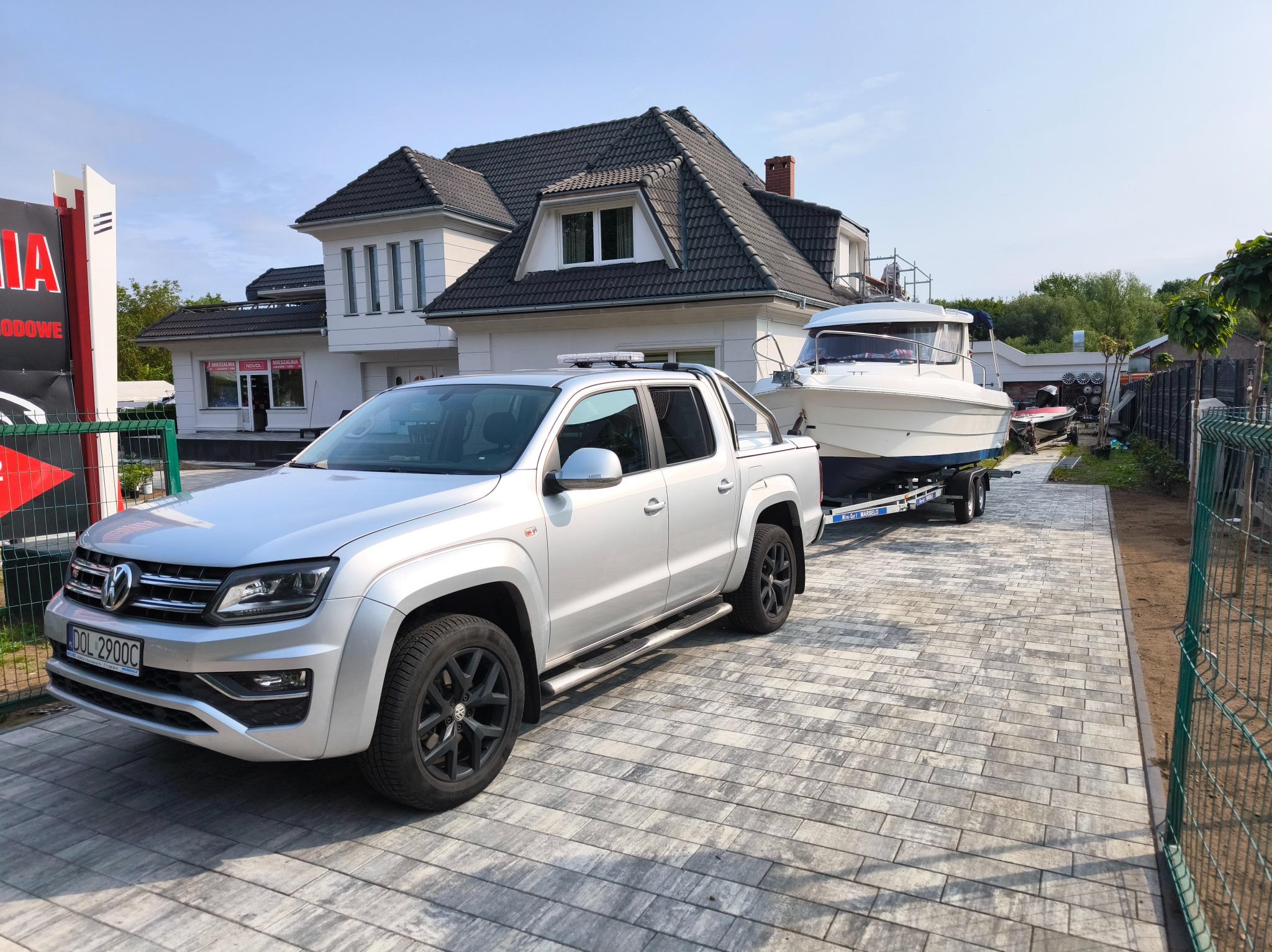 Srebrny pick-up Volkswagen Amarok z przyczepą i łodzią na posesji z kostki brukowej. Widoczny budynek w tle. Transport łodzi.