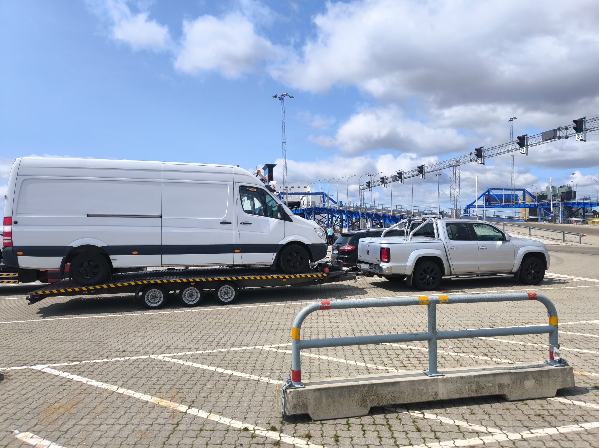 Laweta z białym busem i srebrnym pickupem na parkingu portowym. W tle sygnalizacja świetlna i budynki portowe pod błękitnym niebem z chmurami.