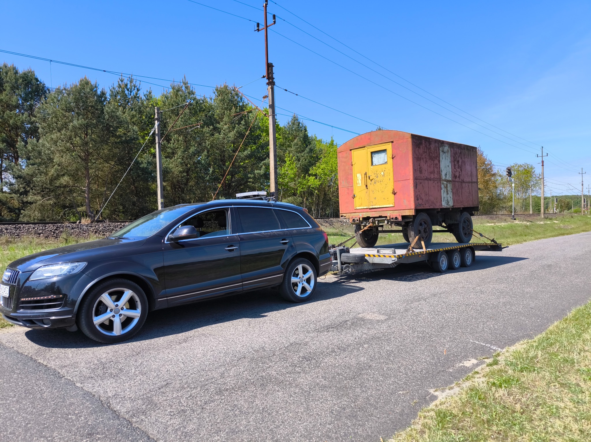 Czarny SUV Audi Q7 ciągnie lawetę z rdzewiejącą, żółto-czerwoną przyczepą budowlaną na drodze z zielonym poboczem i linią kolejową w tle.