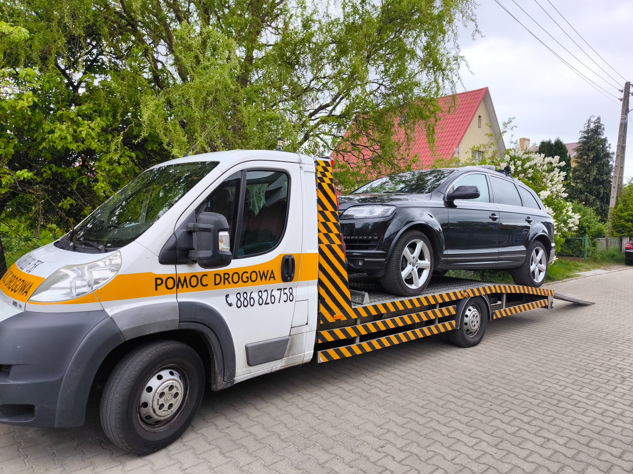 Laweta z czarnym SUV-em Audi Q7 na platformie, zaparkowana na brukowanej drodze, z zielenią w tle. Na lawecie napis 'Pomoc Drogowa' i numer telefonu.