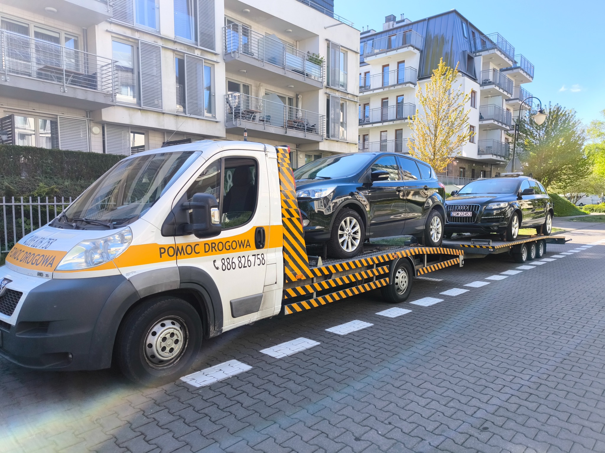 Laweta z dwoma samochodami osobowymi na platformie, zaparkowana na tle nowoczesnego budynku mieszkalnego w słoneczny dzień. Widoczny numer telefonu.