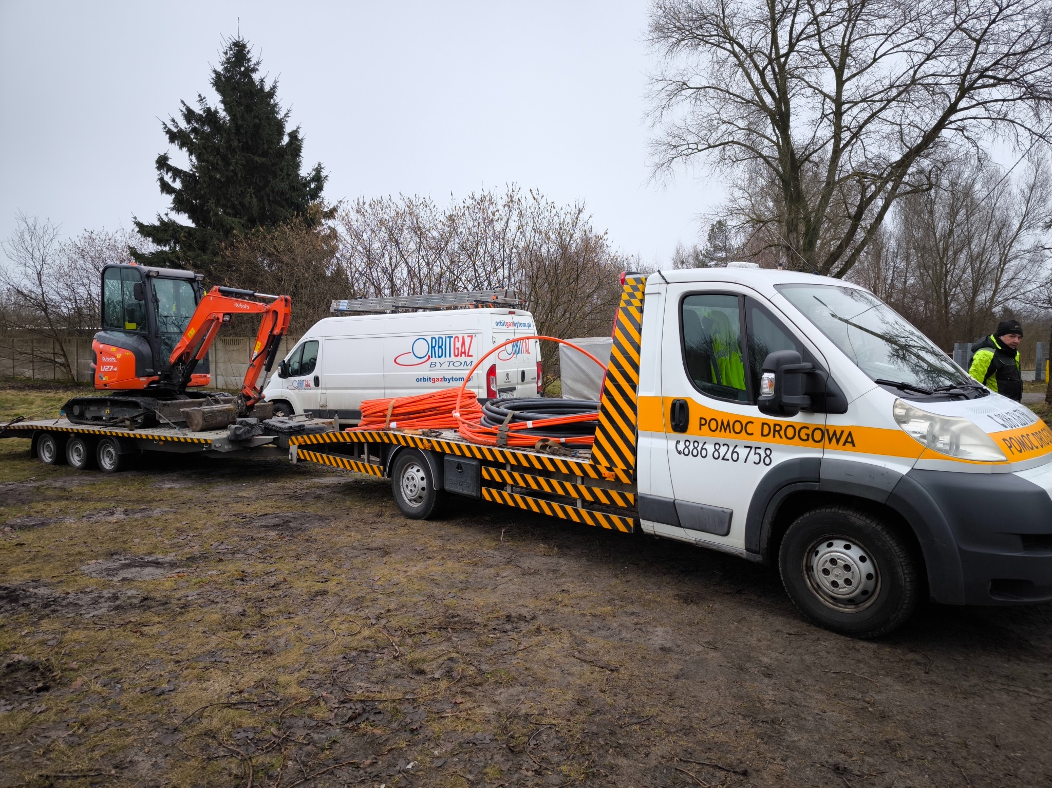 Laweta z minikoparką Kubota i rurami na przyczepie, obok biały bus OrbitGaz Bytom. Pomoc drogowa, teren błotnisty, pochmurny dzień.