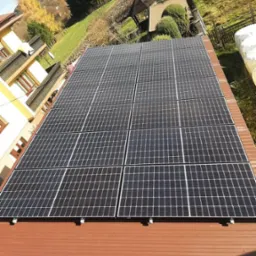 Panel słoneczny na brązowym dachu domu w słoneczny dzień. Widok z g&oacute;ry na panele fotowoltaiczne, dom z ogrodem i pag&oacute;rkowaty krajobraz w tle.