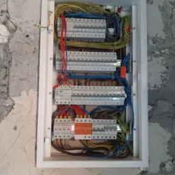 Rozdzielnica elektryczna w białej obudowie, widoczne zabezpieczenia, przewody w różnych kolorach, zamontowana na surowej ścianie.