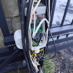 Krzysztof Burak - Otwarta obudowa sterownika bramy przesuwnej z widoczną elektroniką, przewodami i elementami montażowymi. Fragment bramy w tle.