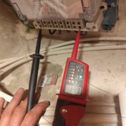 Krzysztof Burak - Pomiar napięcia na liczniku elektrycznym za pomocą próbnika napięcia. Widoczna dłoń elektryka i przewody. Próba odczytu wartości napięcia.