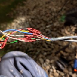 Krzysztof Burak - Połączenie kabli telekomunikacyjnych z użyciem termokurczliwych złączek, widoczne narzędzia i fragment ubrania osoby wykonującej prace na tle żwirowego podłoża.