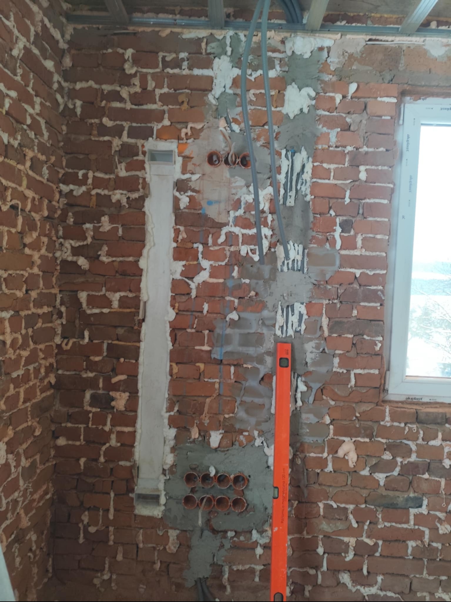 Murowana ściana w trakcie prac instalacyjnych: kable, peszle, poziomica. Fragment ściany z widocznym oknem. Etap prac przed wykończeniem.