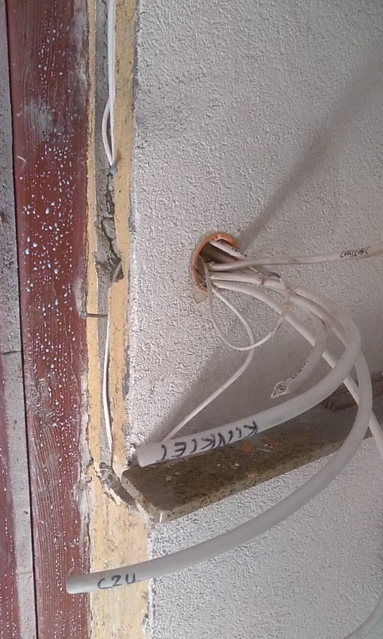 Instalacja elektryczna w trakcie prac: białe przewody wystają z pomarańczowej puszki w ścianie, widoczne napisy na przewodach i fragment drewnianej ramy drzwi.
