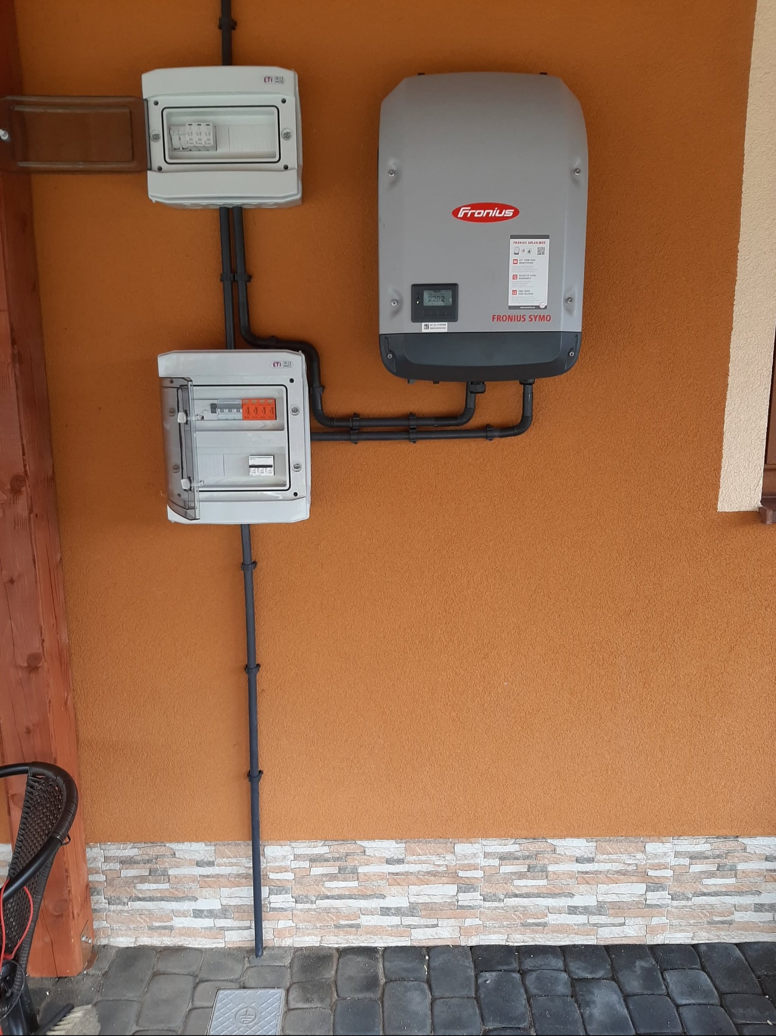 Zewnętrzna instalacja fotowoltaiczna na pomarańczowej ścianie budynku. Widoczne dwie skrzynki rozdzielcze ETI i falownik Fronius Symo, połączone czarnymi rurkami instalacyjnymi.