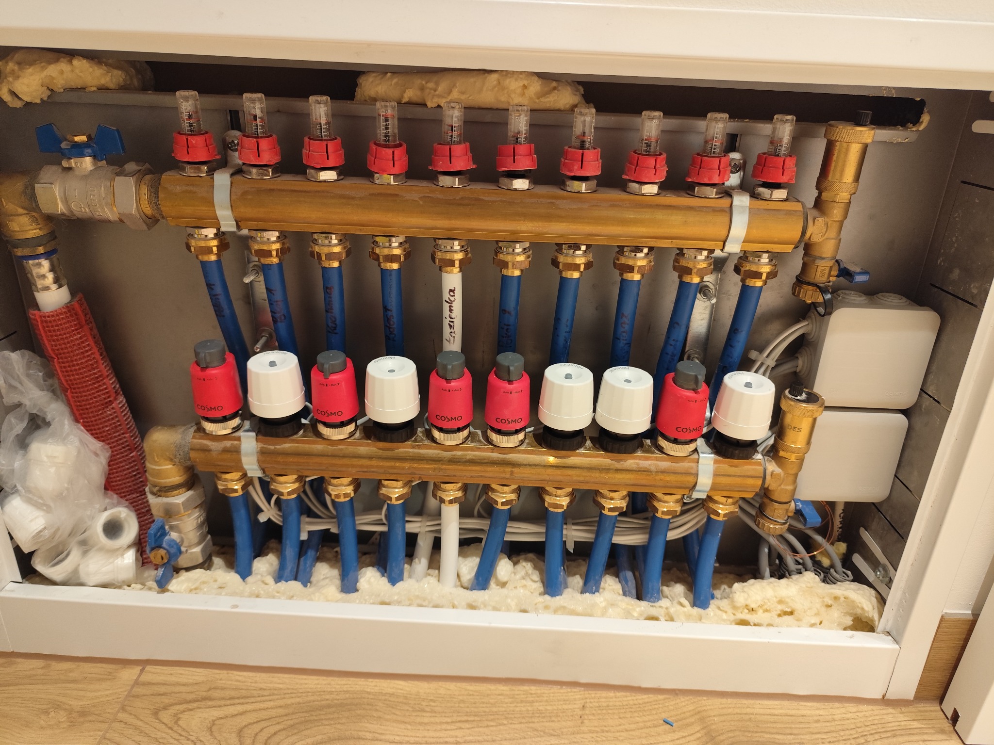 Rozdzielacz ogrzewania podłogowego w białej szafce, z rurami PEX w kolorze niebieskim, z izolacją piankową i elementami regulacyjnymi Cosmo. Widok z bliska.