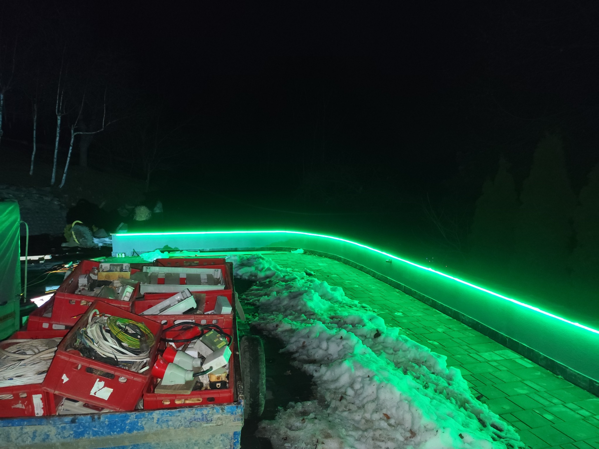 Zimowy krajobraz nocą z zielonym oświetleniem LED wzdłuż ścieżki i skrzynkami z kablami, sugerujący prace elektryczne na zewnątrz.