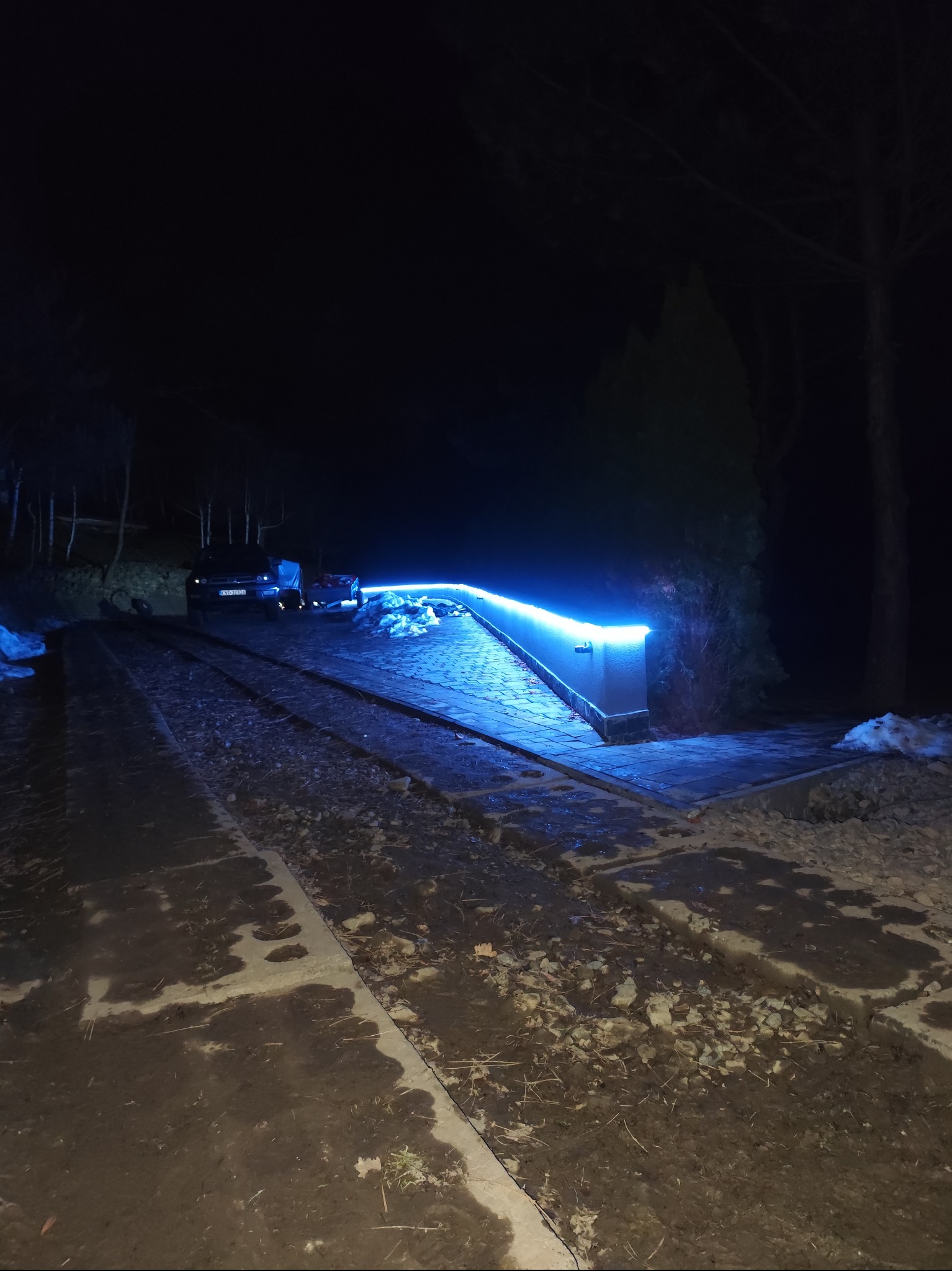 Nocne ujęcie podjazdu z niebieskim oświetleniem LED, samochód w tle. Zimowa sceneria z resztkami śniegu. Nietypowe oświetlenie podjazdu.