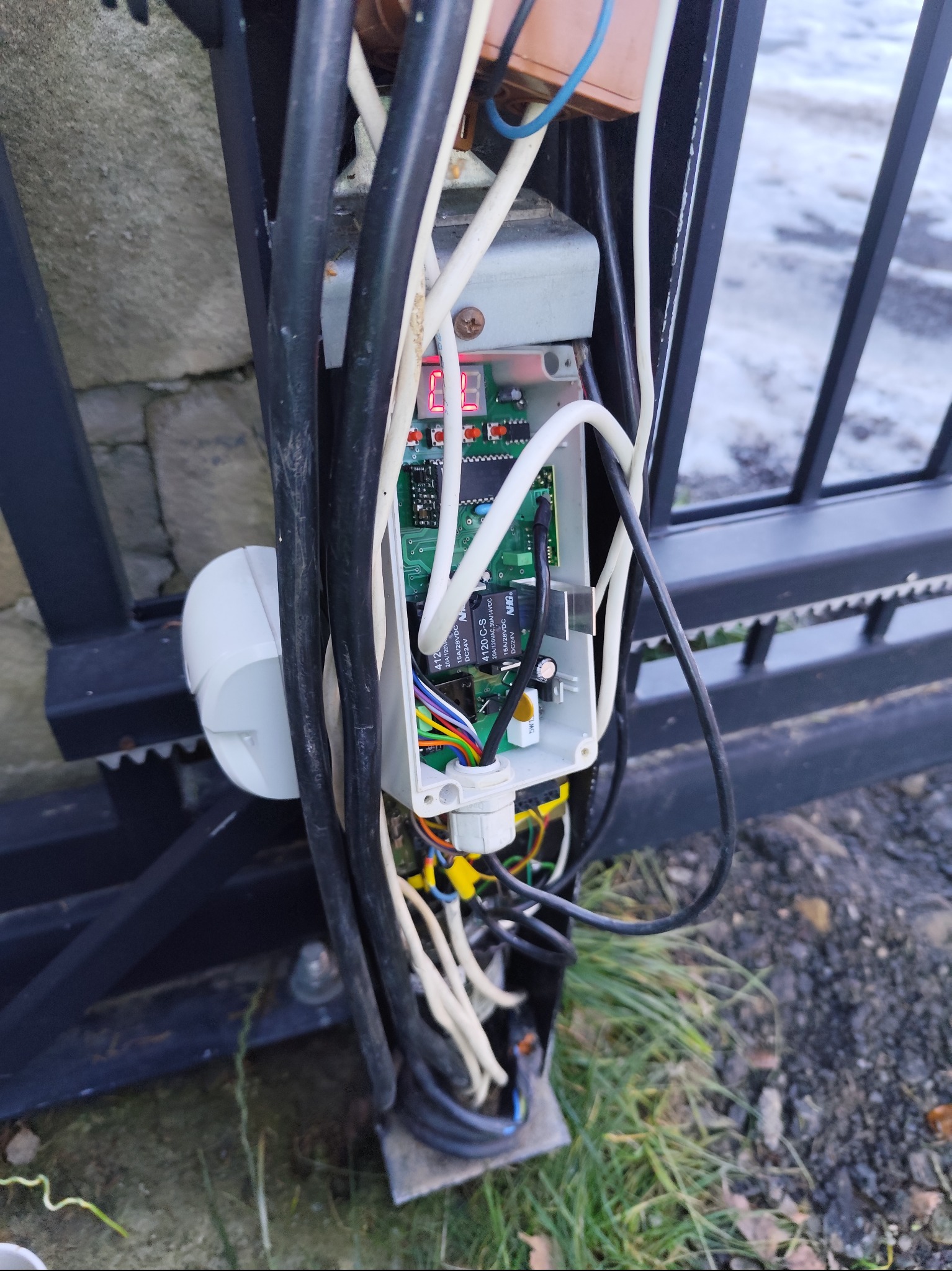 Otwarta obudowa sterownika bramy przesuwnej z widoczną elektroniką, przewodami i elementami montażowymi. Fragment bramy w tle.