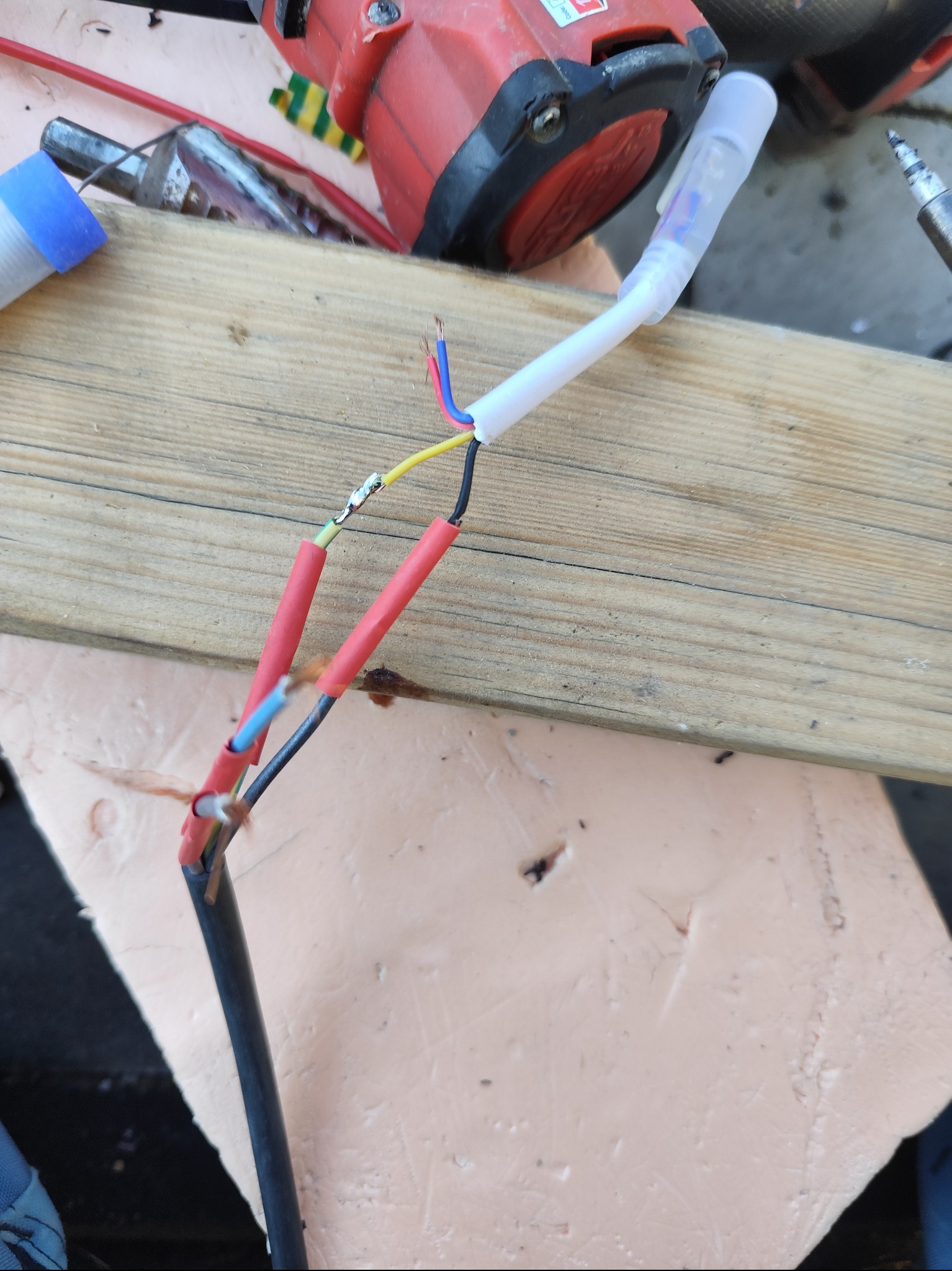 Połączenie przewodów elektrycznych z użyciem koszulek termokurczliwych i lutownicy na drewnianej desce. Widoczne kolory przewodów: niebieski, żółty, czarny.