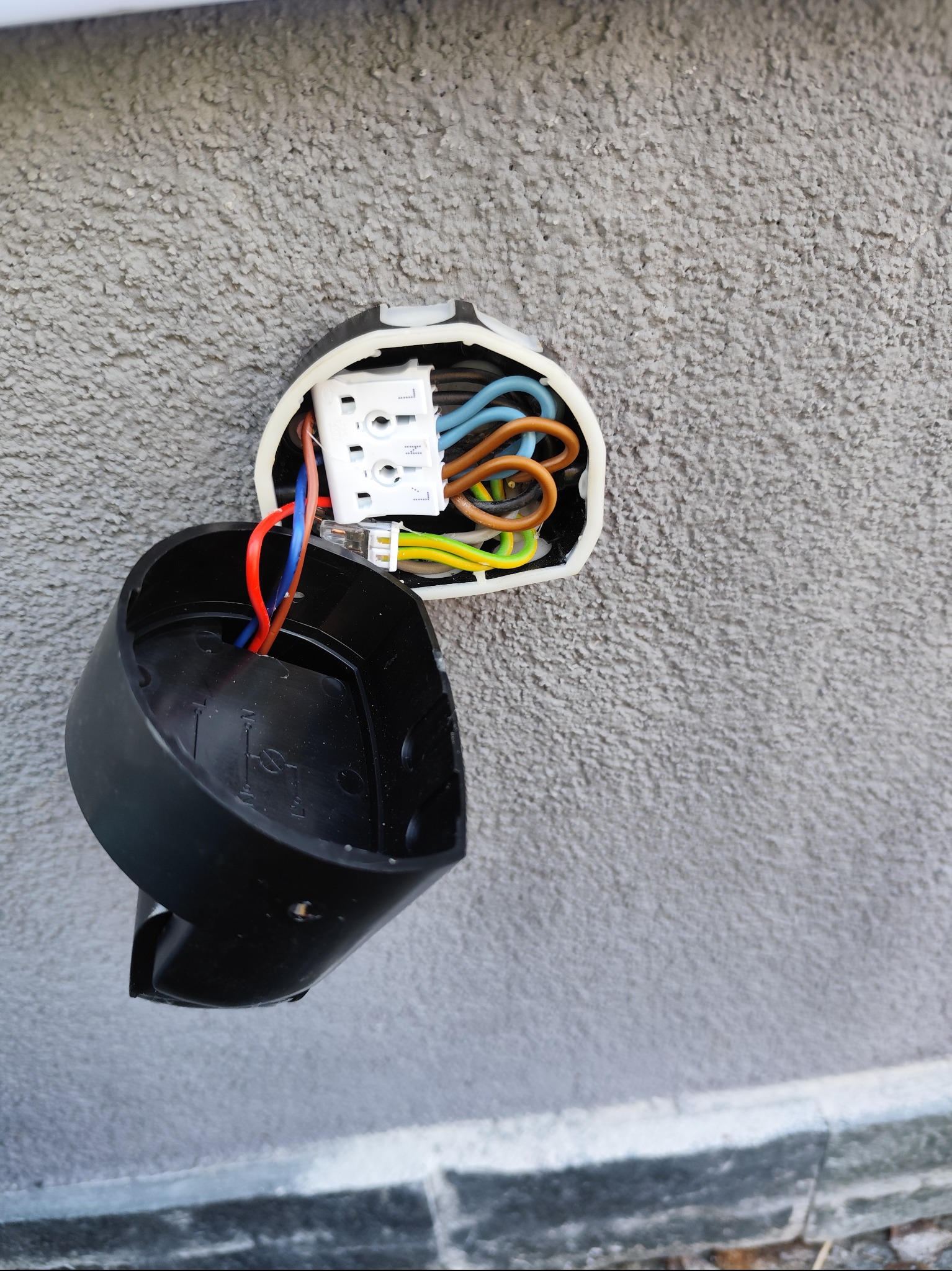 Otwarta puszka elektryczna na szarej, chropowatej ścianie zewnętrznej. Widoczne kolorowe przewody i kostka. Czarna obudowa puszki zwisa luźno.