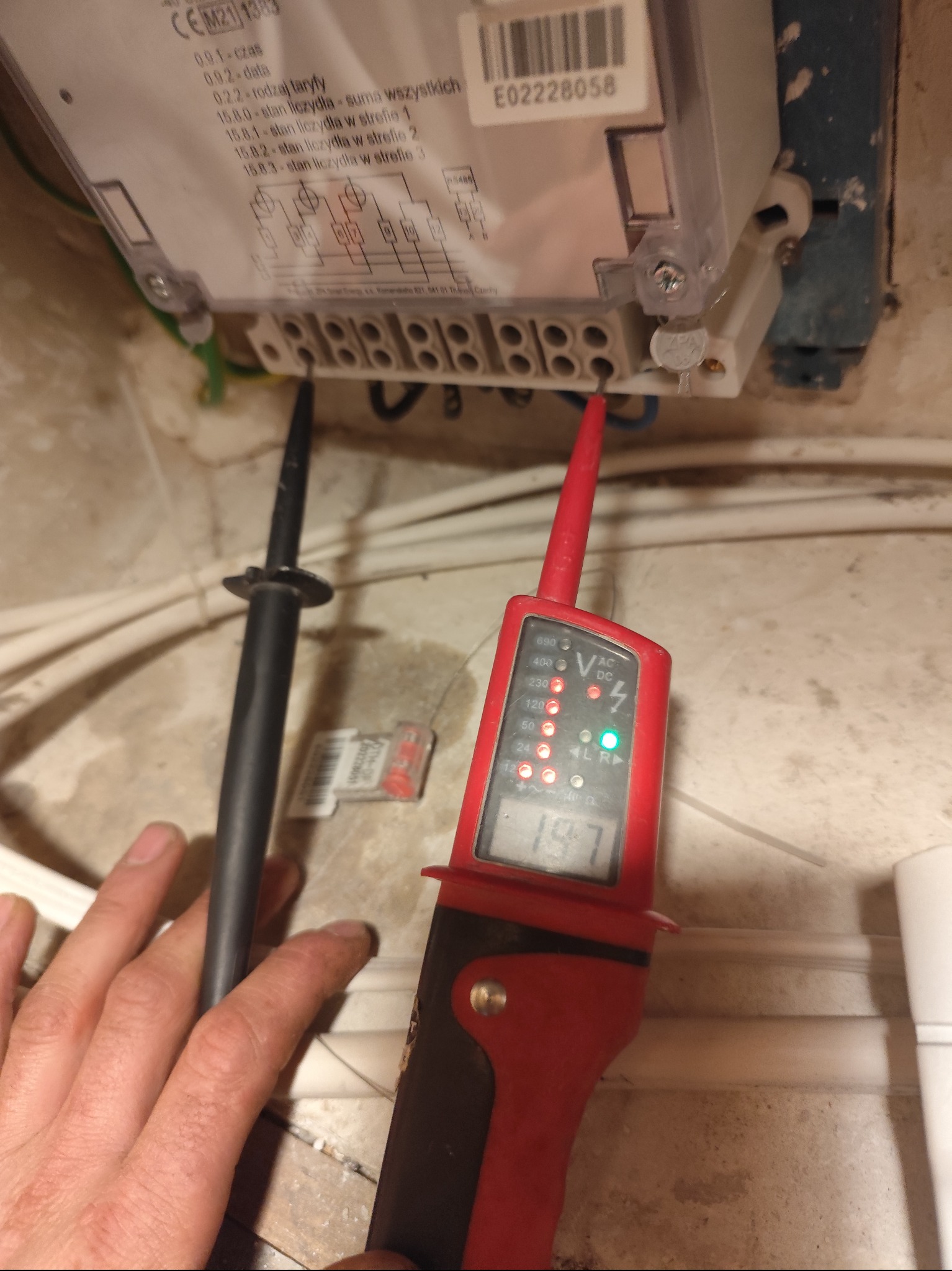 Pomiar napięcia na liczniku elektrycznym za pomocą próbnika napięcia. Widoczna dłoń elektryka i przewody. Próba odczytu wartości napięcia.