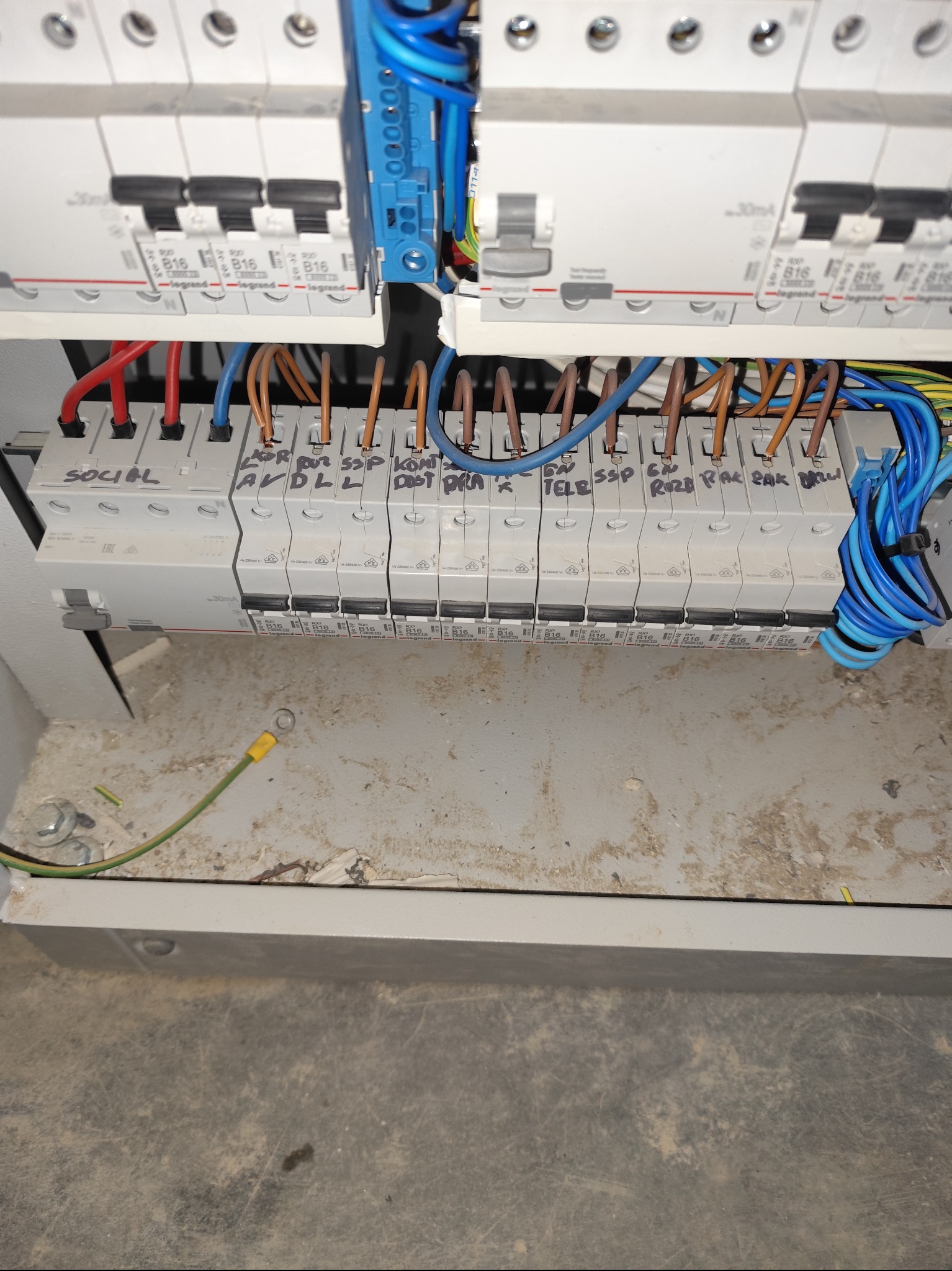 Rozdzielnica elektryczna z bezpiecznikami Legrand, oznaczonymi opisami, z widocznymi przewodami w kolorach niebieskim, brązowym i czerwonym. Widoczne ślady użytkowania.