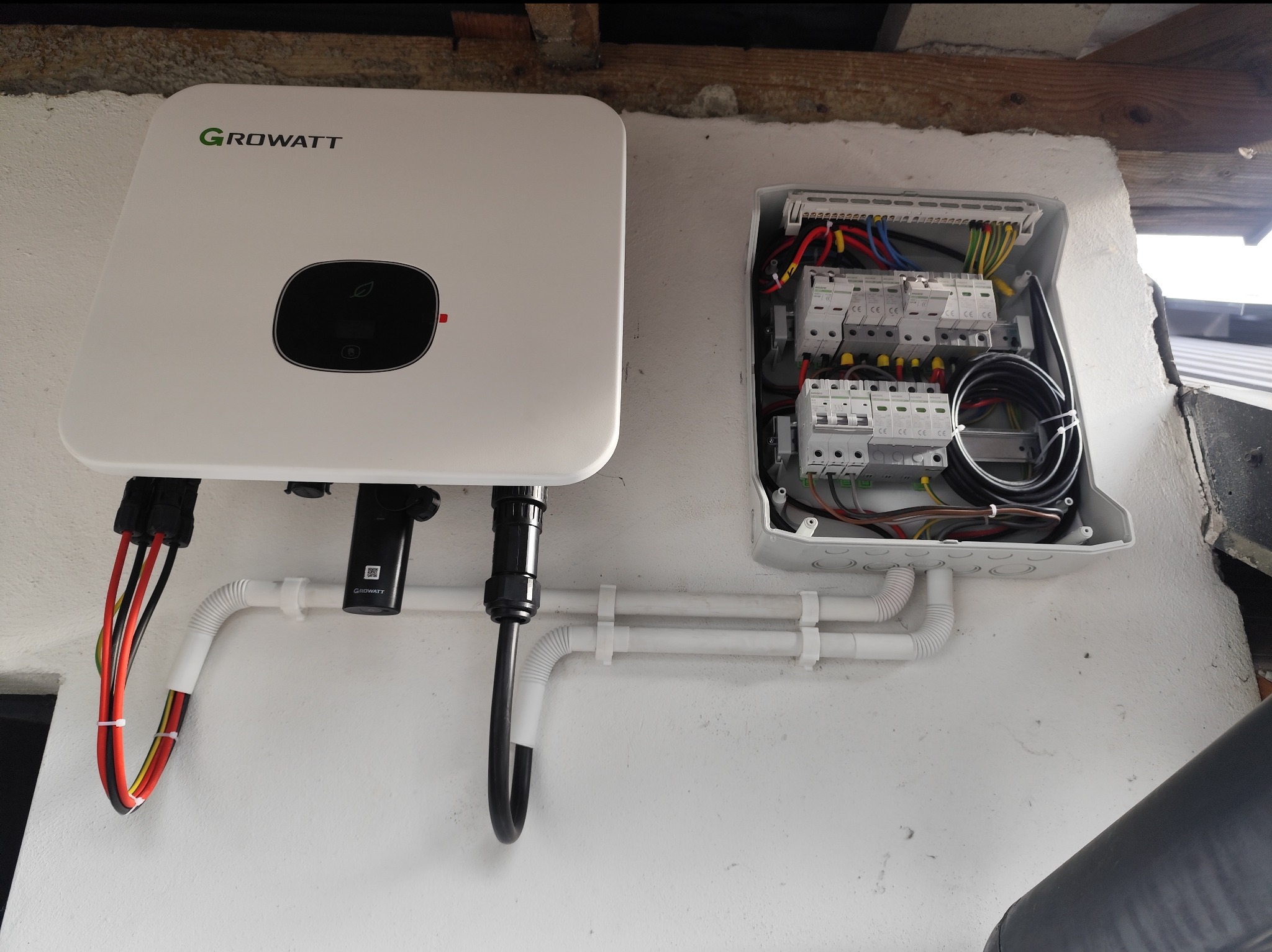 Biały inwerter Growatt zamontowany na ścianie obok otwartej skrzynki z bezpiecznikami i przewodami. Widoczne rurki ochronne i kable.