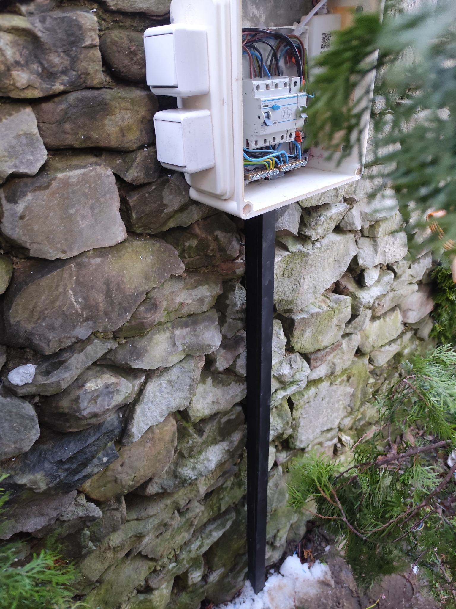 Biała skrzynka elektryczna z bezpiecznikami, zamontowana na czarnym słupku przy kamiennym murze, z fragmentem zielonego krzewu w tle.