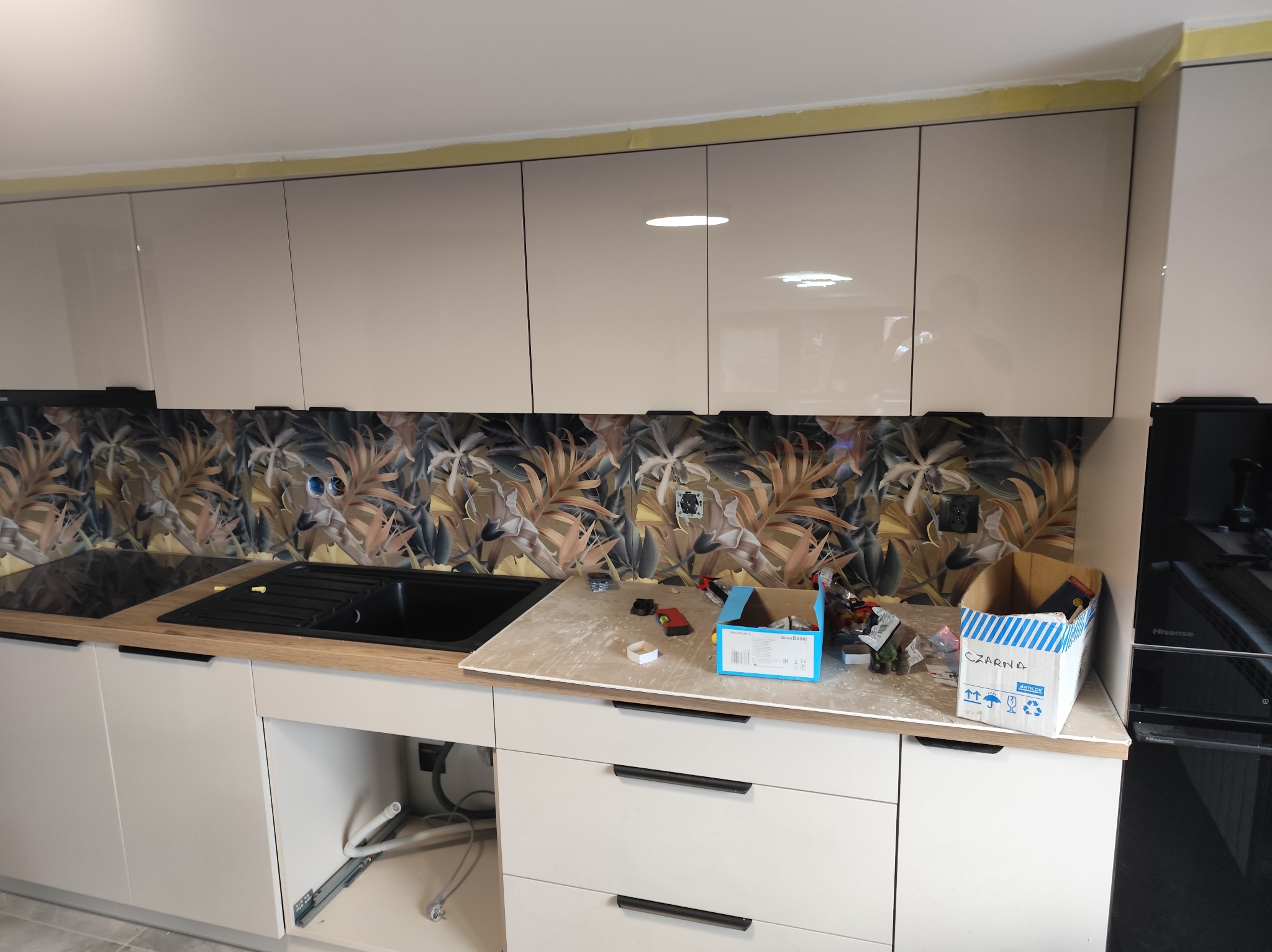 Montaż kuchni: beżowe szafki z czarnymi uchwytami, wzorzysty panel nad blatem, widoczne narzędzia i akcesoria montażowe. Etap prac wykończeniowych.