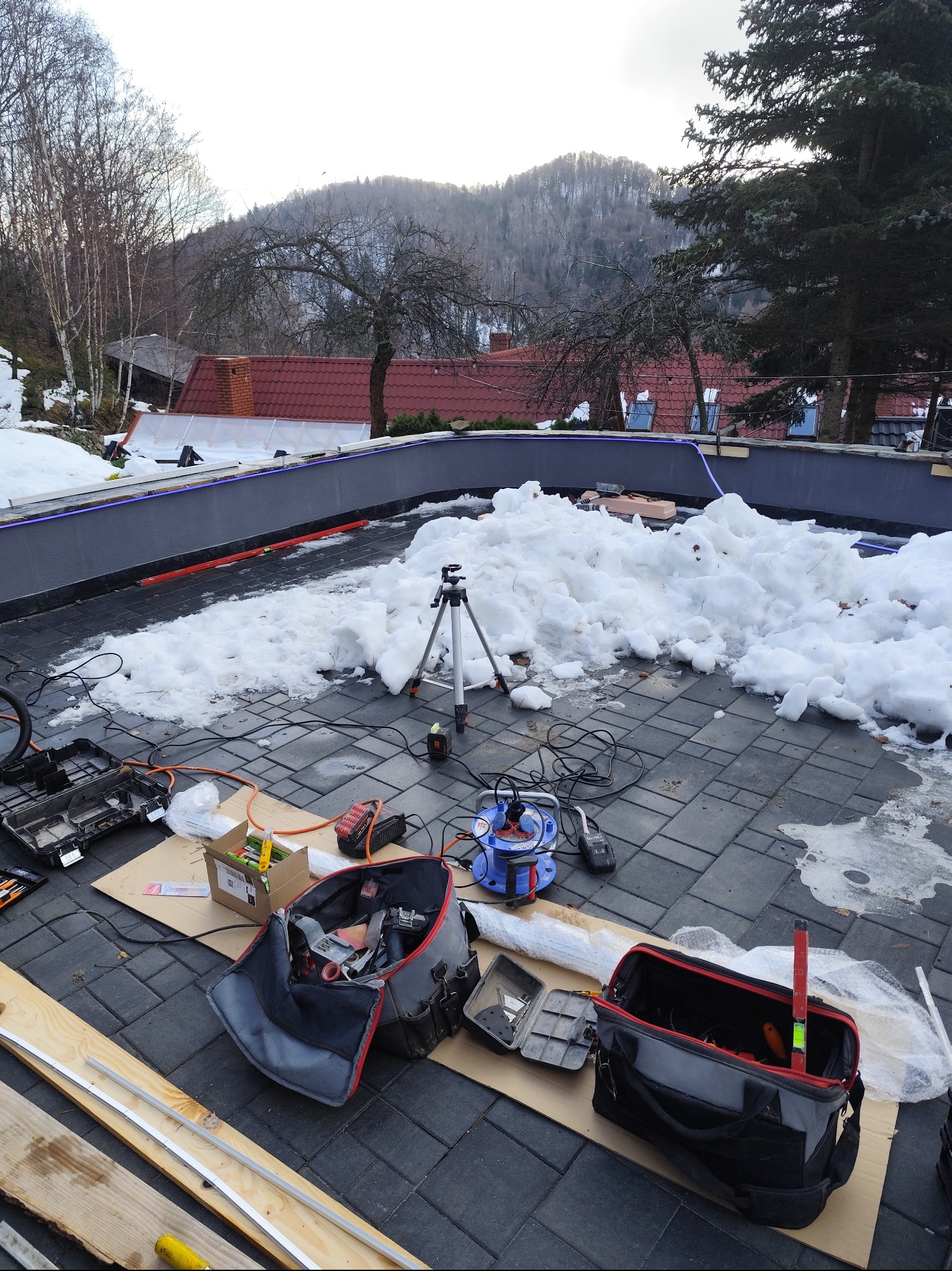 Montaż oświetlenia zewnętrznego na tarasie z kostki brukowej w zimowej scenerii. Widoczne narzędzia, torby monterskie i statyw. W tle góry i budynki.