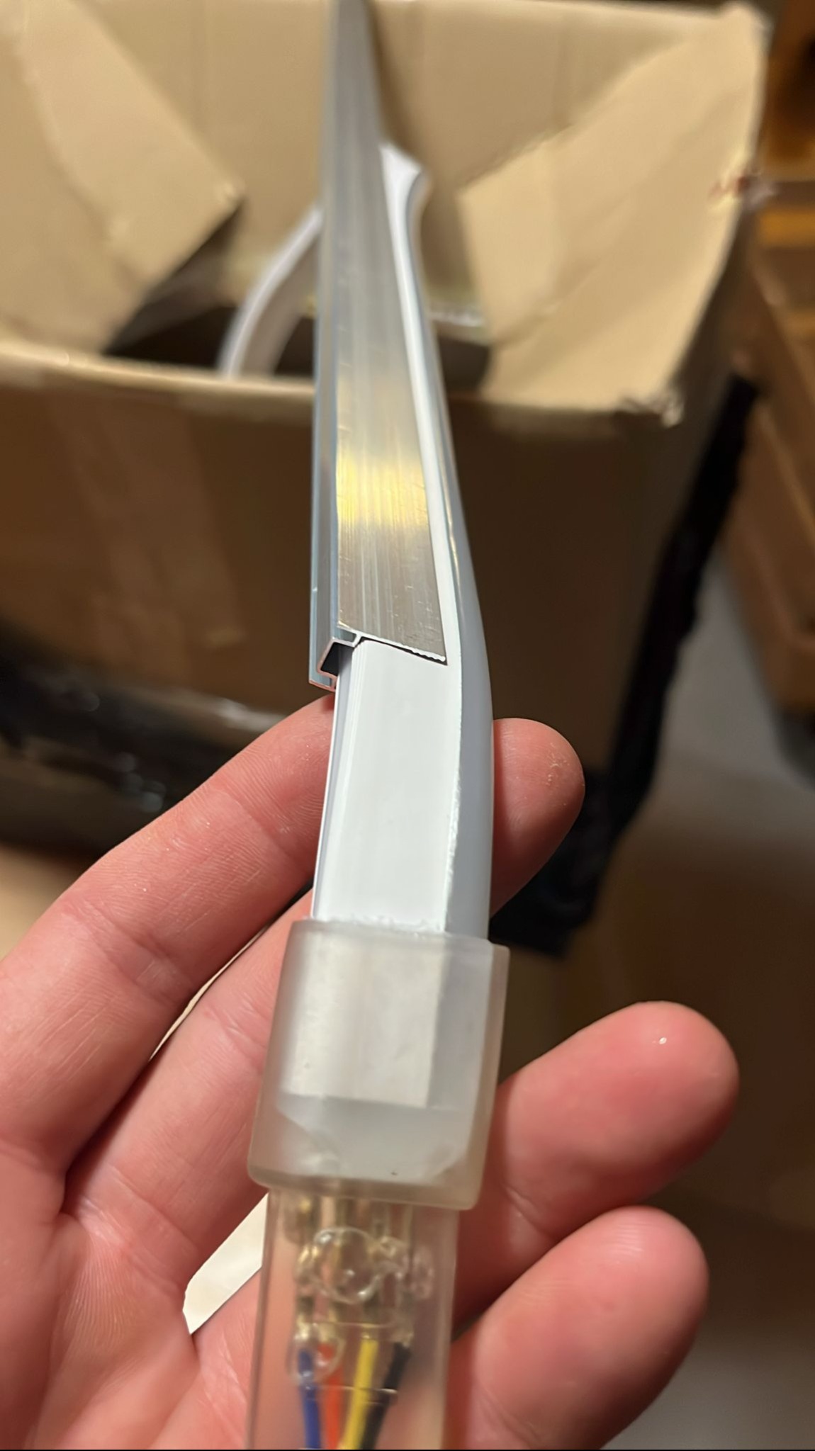 Profil LED z widocznymi przewodami w przezroczystej osłonie, trzymany w dłoni na tle kartonowego pudła. Detal z bliska.
