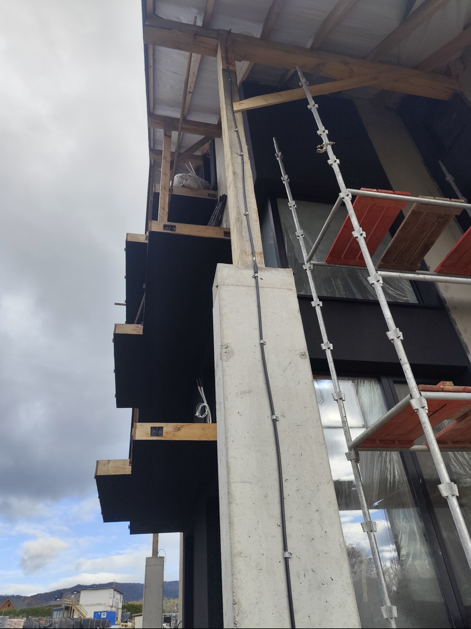 Budynek w trakcie wykończenia z widocznymi rusztowaniami, elementami drewnianymi i instalacją elektryczną na betonowym słupie. Ujęcie z dołu, pochmurne niebo.