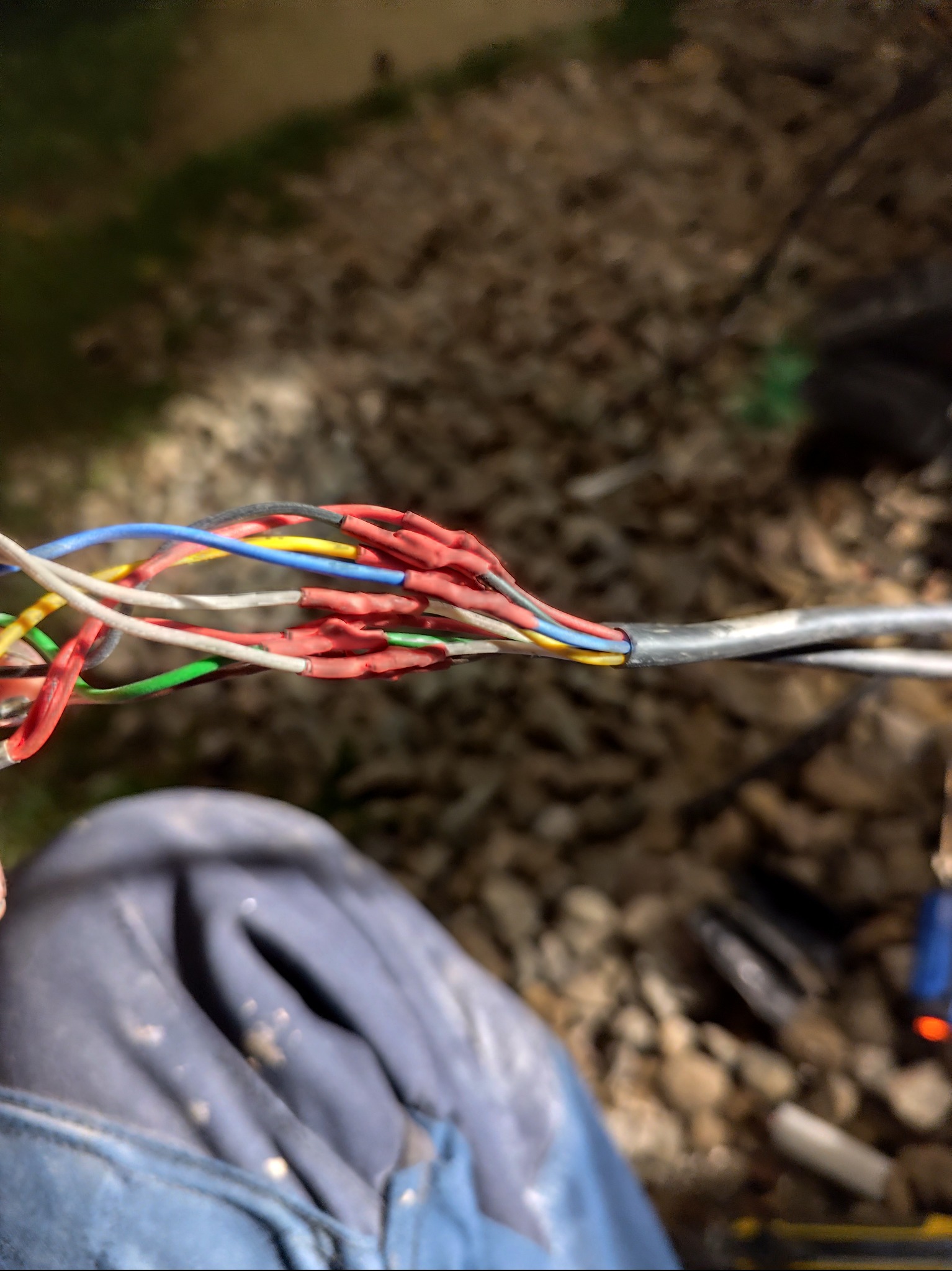 Połączenie kabli telekomunikacyjnych z użyciem termokurczliwych złączek, widoczne narzędzia i fragment ubrania osoby wykonującej prace na tle żwirowego podłoża.