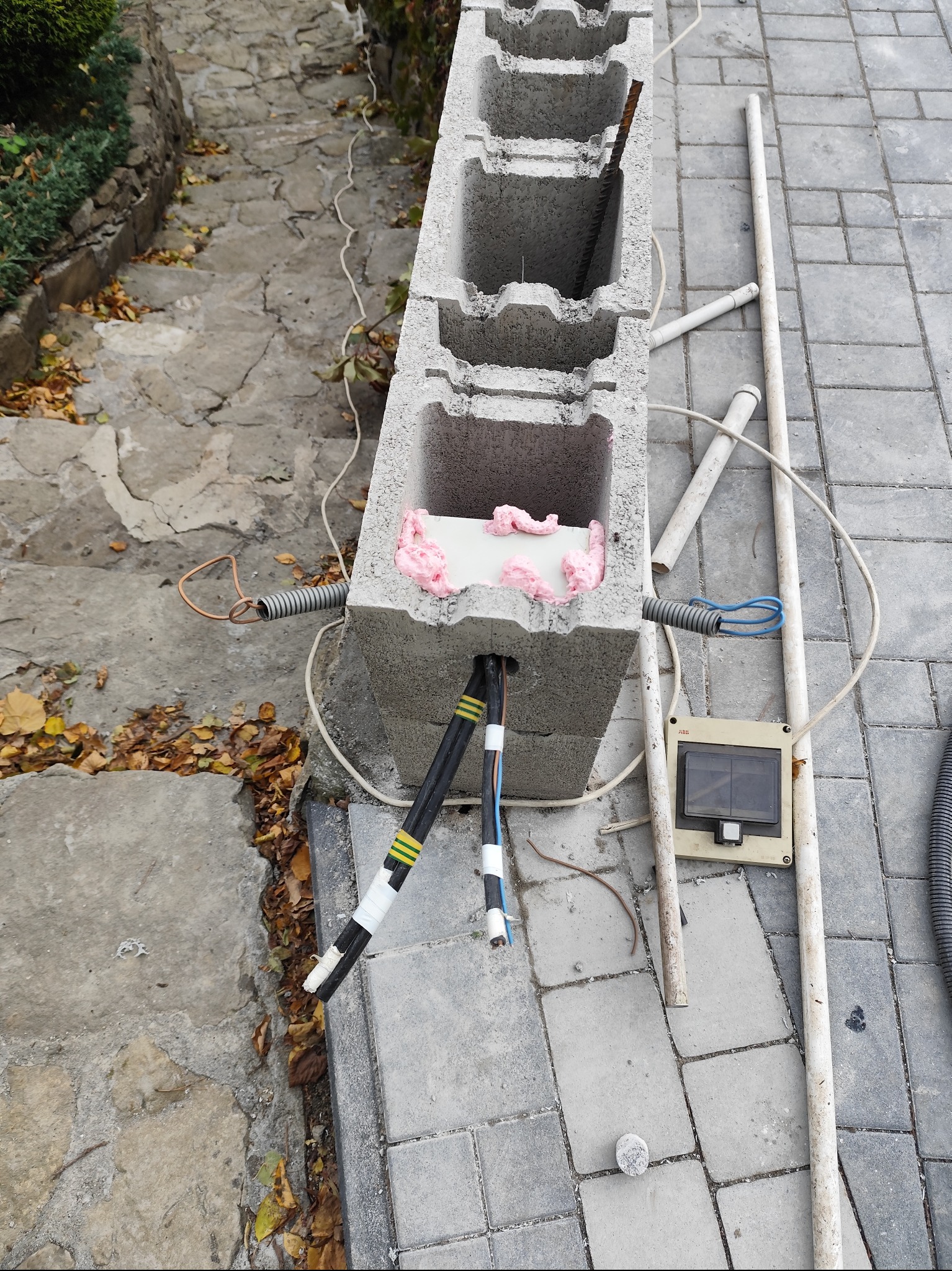 Betonowy pustak z przewodami elektrycznymi i pianką montażową, skrzynka elektryczna obok. Widoczne kable w peszlach i rury PCV. Prace instalacyjne na zewnątrz budynku.