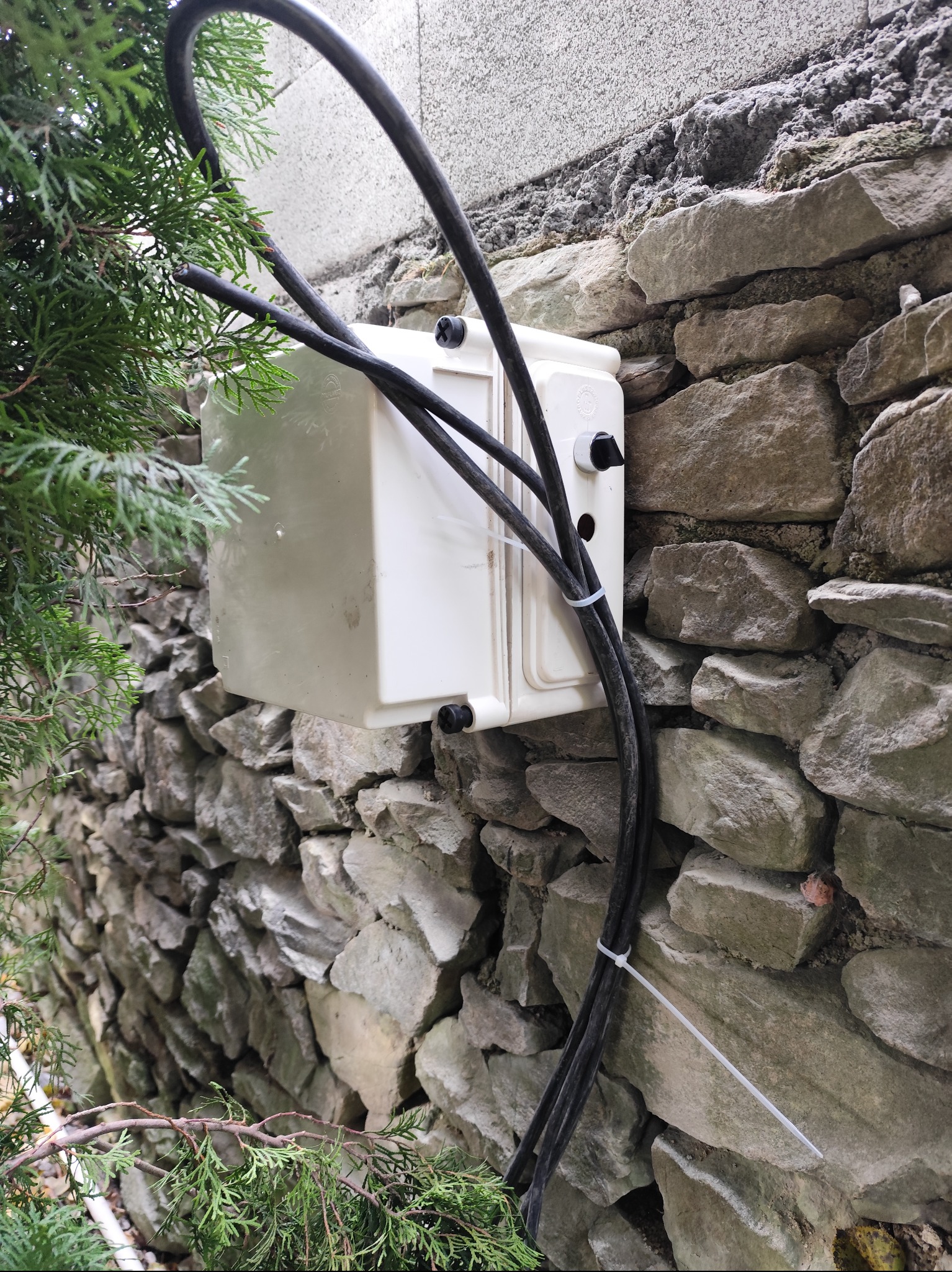 Biała skrzynka elektryczna z czarnymi kablami przymocowana do kamiennej ściany zewnętrznej, zabezpieczona opaskami zaciskowymi, z fragmentem zielonej roślinności po lewej.