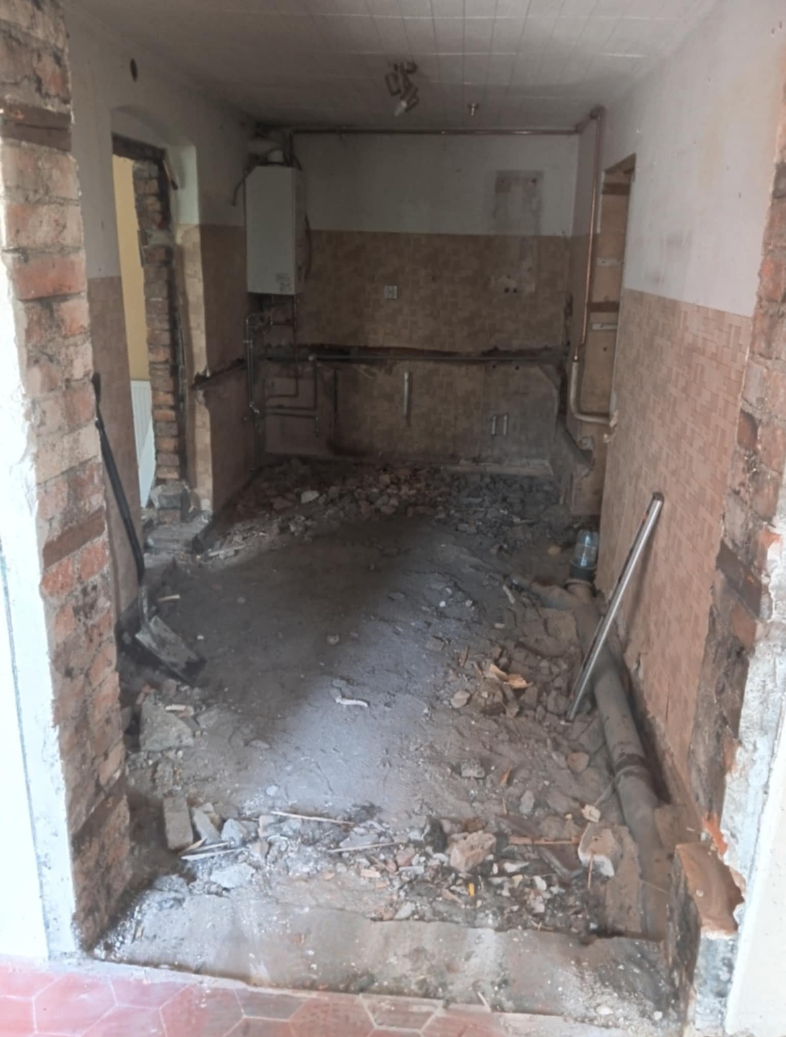 Demontaż starej łazienki: gruz, odsłonięte cegły, instalacje. Widok na wnętrze w trakcie prac, z fragmentami płytek i rur. Kotłownia w tle.