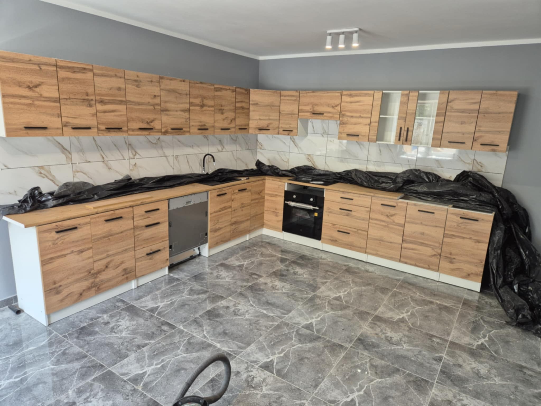 Jasna kuchnia z drewnianymi frontami i marmurowymi płytkami, częściowo zakryta folią ochronną, w trakcie montażu.