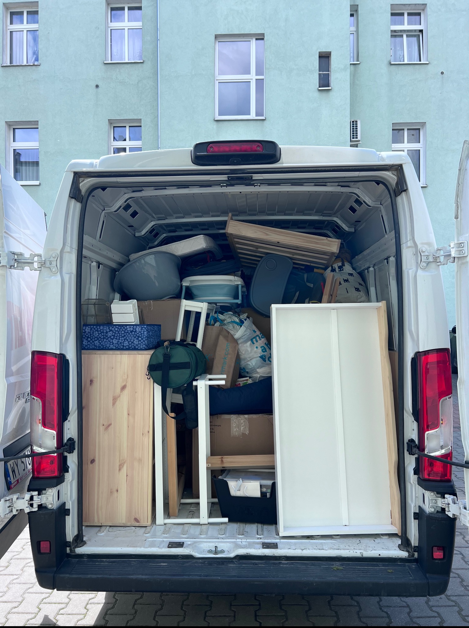 Otwarty bagażnik białego busa wypełniony różnymi przedmiotami, w tle budynek z błękitną elewacją i oknami. Widok z tyłu pojazdu.