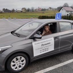 OSK Dynamic - Szary samochód szkoły nauki jazdy z literą 'L' na dachu, zaparkowany na placu. Za nim budynki mieszkalne i zielone wzgórze. W środku siedzi blondynka.
