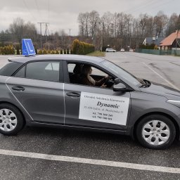 OSK Dynamic - Szary samochód z niebieskim znakiem 'L' na dachu, zaparkowany na placu manewrowym. Na drzwiach naklejka z nazwą ośrodka szkolenia kierowców 'Dynamic'.
