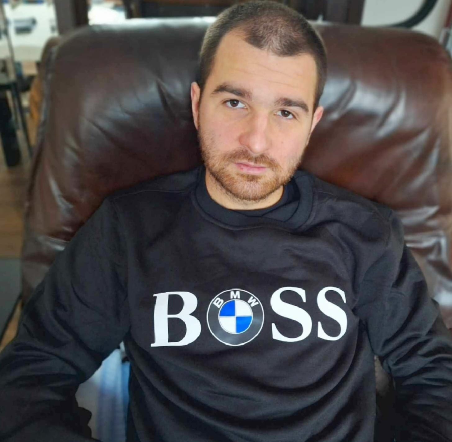 Młody mężczyzna w czarnej bluzie z logo BMW BOSS, siedzący w skórzanym fotelu. Ujęcie od przodu, skupione na twarzy i górnej części ciała.