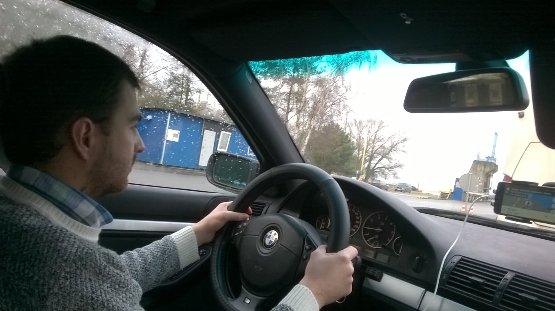 Mężczyzna w swetrze prowadzi BMW; widok z wnętrza auta na mokrą drogę, budynek i drzewa. Smartfon z nawigacją na desce rozdzielczej.