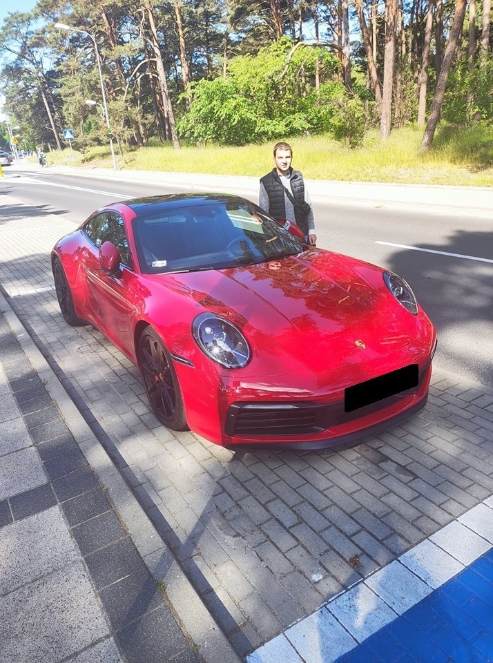 Czerwone Porsche 911 zaparkowane na chodniku, obok stoi mężczyzna. Drzewa i zieleń w tle. Lekko pochylony kadr, widoczny cień samochodu na kostce.