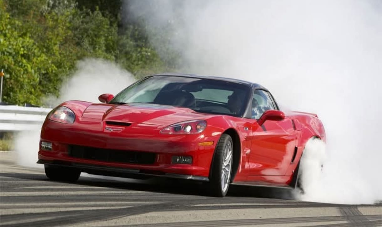 Czerwony sportowy samochód w poślizgu, z dymiącymi oponami na asfalcie, dynamiczne ujęcie z perspektywy bocznej, zielone drzewa w tle.