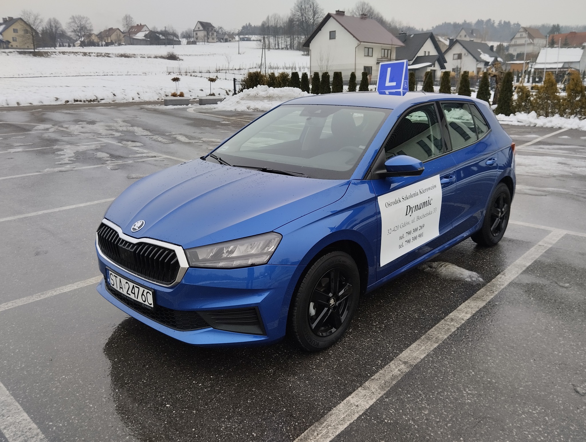 Niebieska Skoda z oznaczeniem 'L' na dachu, zaparkowana na mokrym parkingu. Na drzwiach naklejka z nazwą ośrodka szkolenia kierowców 'Dynamic'. Zimowy krajobraz w tle.