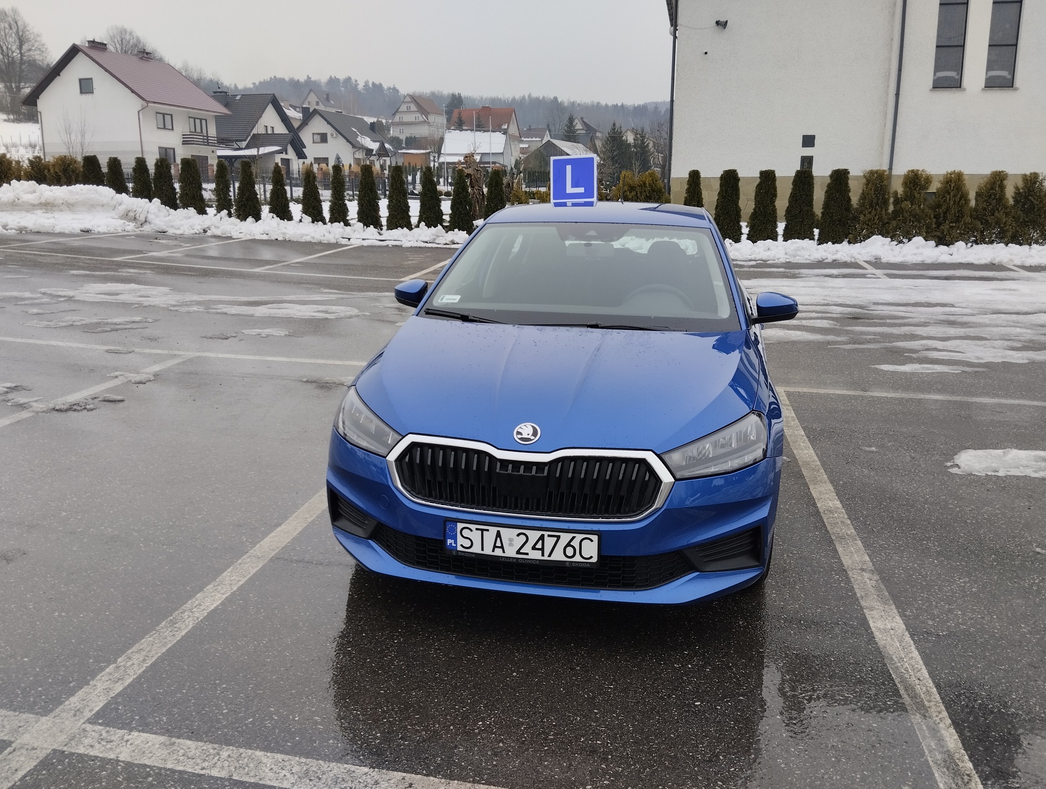 Niebieska Skoda z literą 'L' na dachu, zaparkowana na mokrym parkingu. W tle budynki mieszkalne i drzewa. Tablica rejestracyjna STA 2476C.