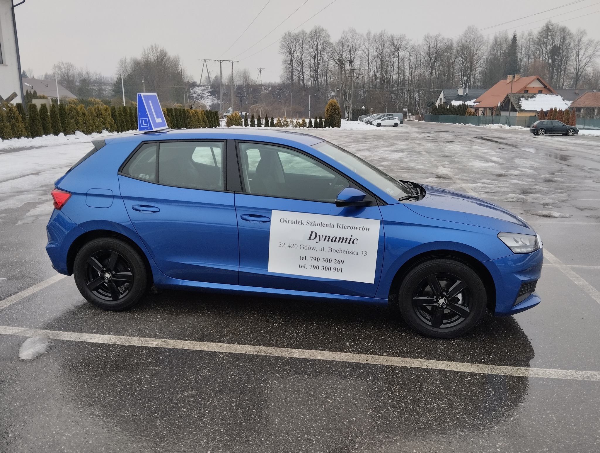 Niebieski samochód z oznaczeniem 'L' na dachu, zaparkowany na parkingu. Na drzwiach naklejka z nazwą i danymi kontaktowymi ośrodka szkolenia kierowców 'Dynamic'.