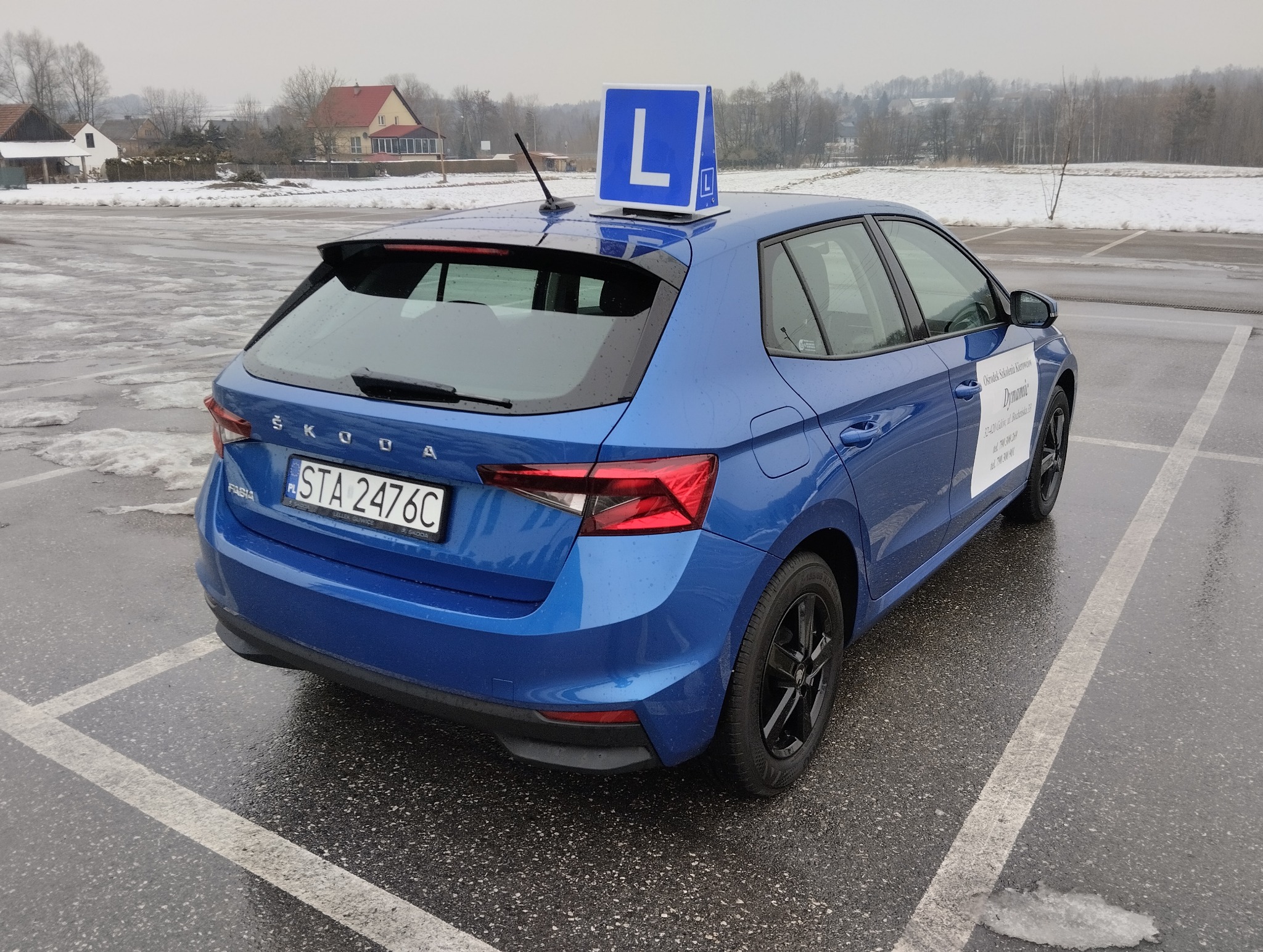 Niebieska Skoda Fabia z literą 'L' na dachu, zaparkowana na mokrym placu. Widoczny tył pojazdu, tablica rejestracyjna i logo szkoły jazdy na drzwiach.