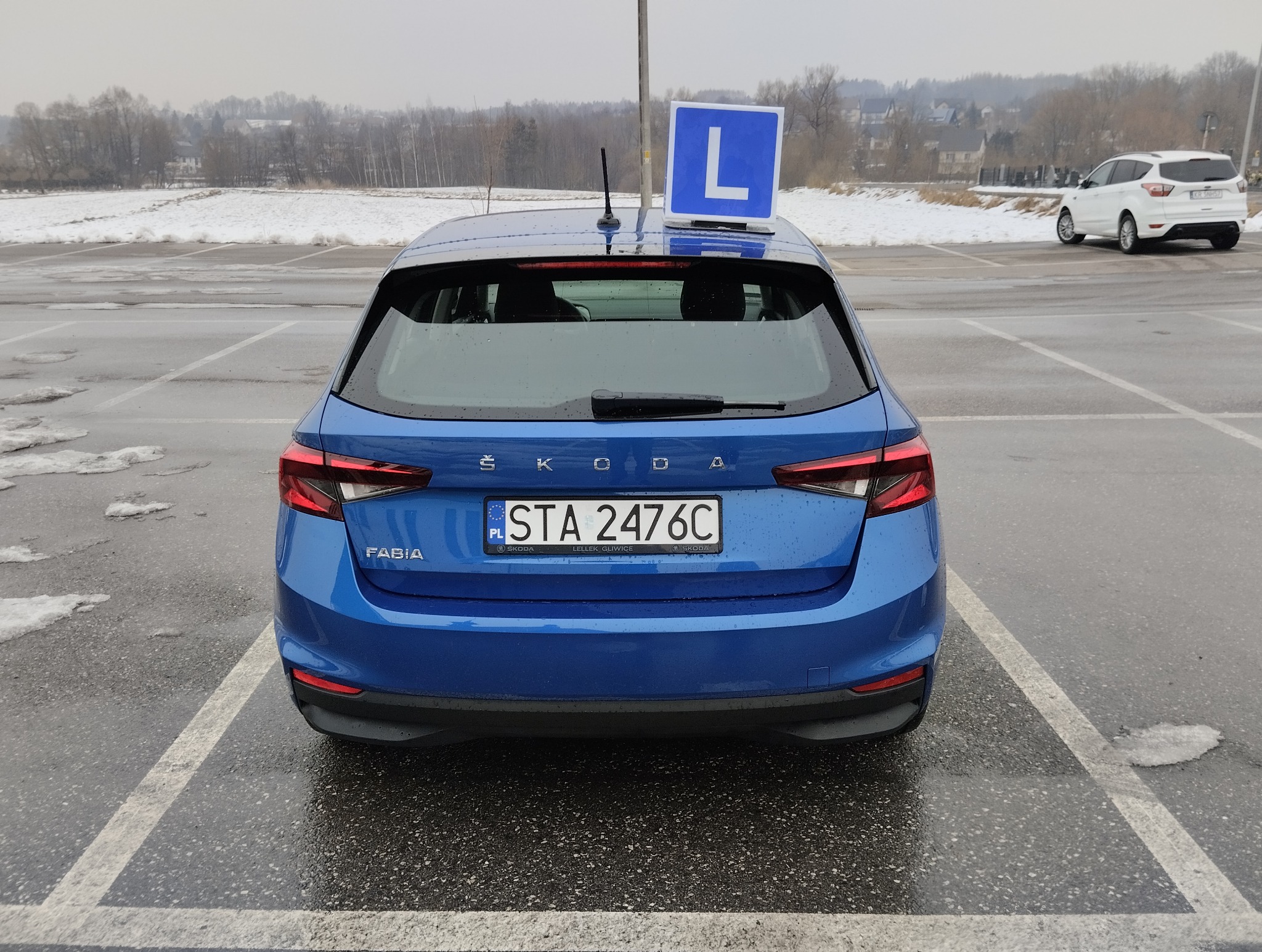 Niebieska Skoda Fabia z symbolem 'L' na dachu, stojąca na parkingu w pochmurny dzień. Widok z tyłu, tablica rejestracyjna STA 2476C. W tle zasnieżone pole i zabudowania.