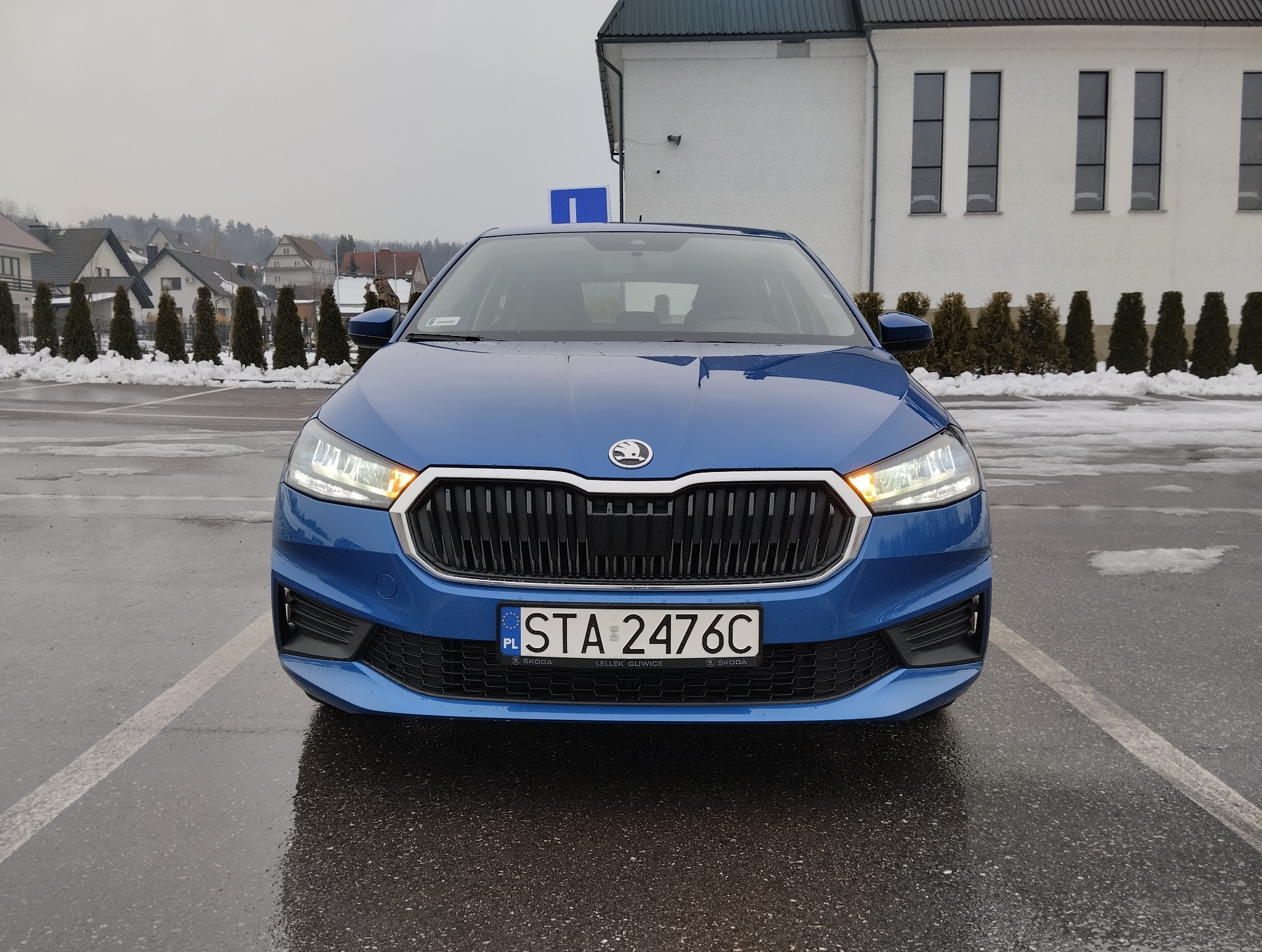 Niebieska Skoda z tablicą rejestracyjną STA 2476C na parkingu szkoły jazdy, widok z przodu, w tle budynek z logo szkoły nauki jazdy.