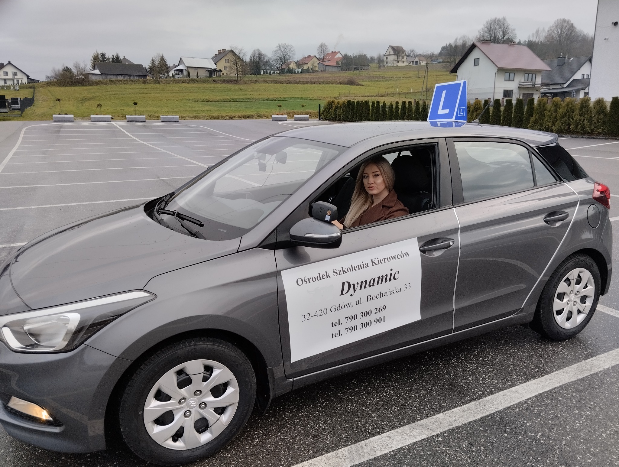Szary samochód szkoły nauki jazdy z literą 'L' na dachu, zaparkowany na placu. Za nim budynki mieszkalne i zielone wzgórze. W środku siedzi blondynka.