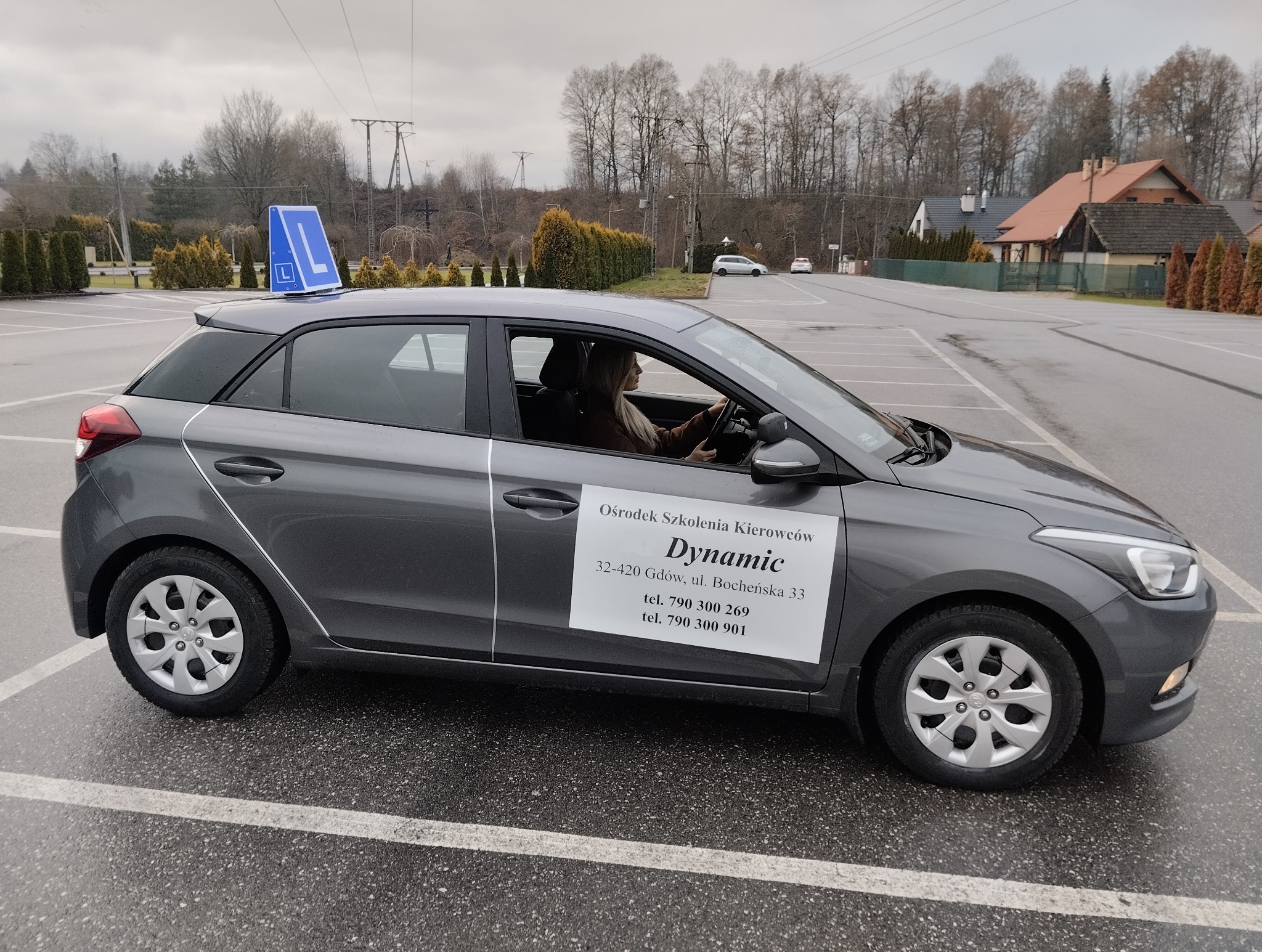 Szary samochód z niebieskim znakiem 'L' na dachu, zaparkowany na placu manewrowym. Na drzwiach naklejka z nazwą ośrodka szkolenia kierowców 'Dynamic'.