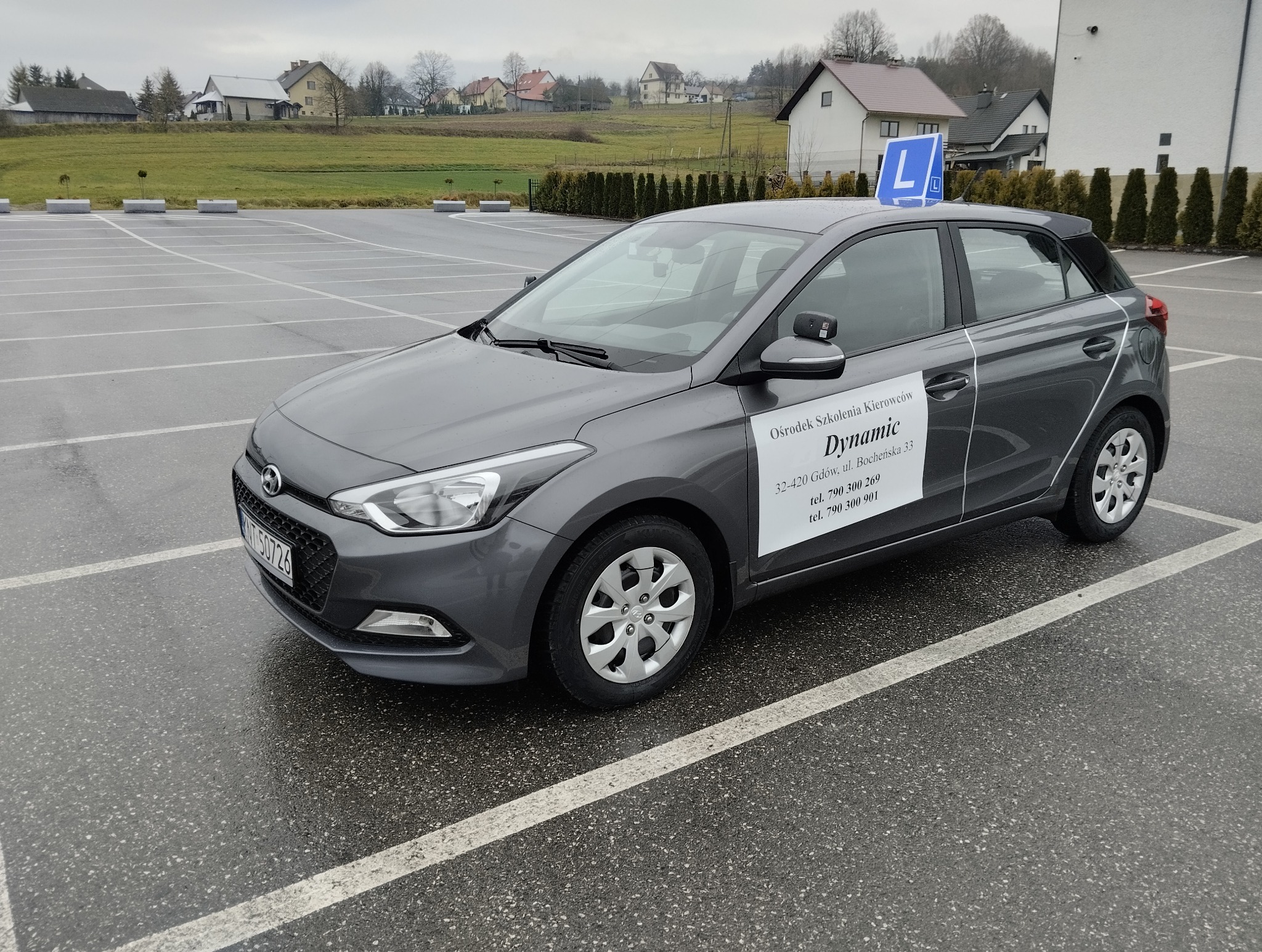 Szary samochód z niebieską literą L na dachu, zaparkowany na mokrym parkingu. Na drzwiach naklejka z nazwą i danymi kontaktowymi ośrodka szkolenia kierowców.
