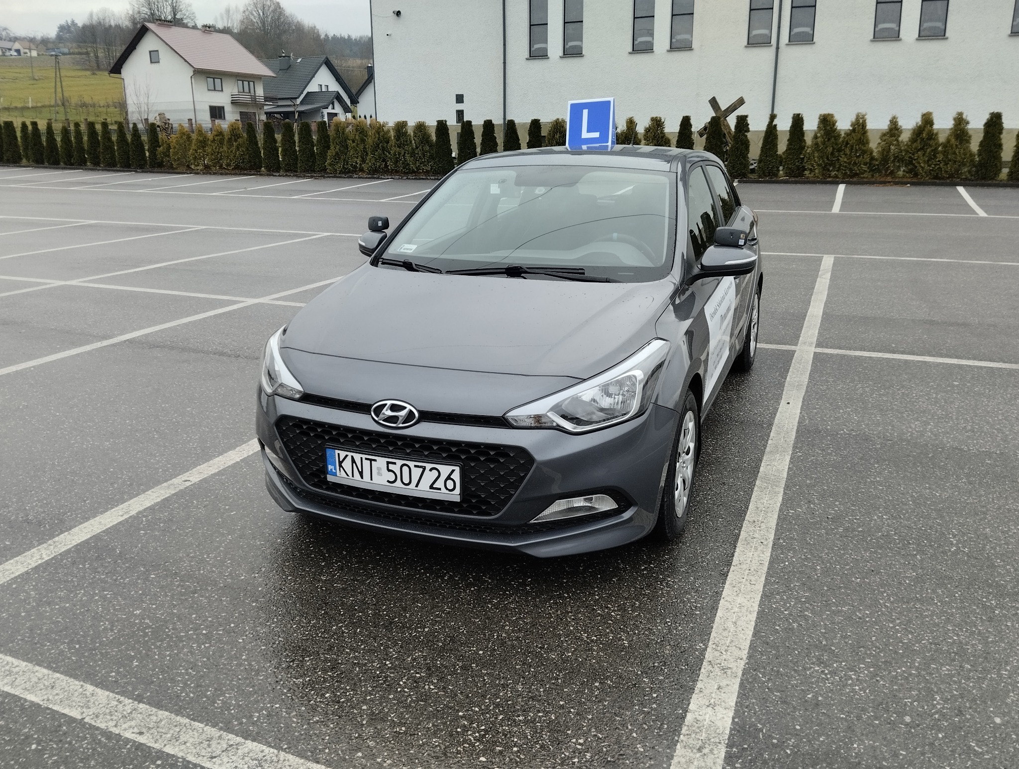 Szary samochód z niebieską literą 'L' na dachu, zaparkowany na mokrym parkingu. Widoczny numer rejestracyjny i budynek w tle. Auto z naklejką firmy.